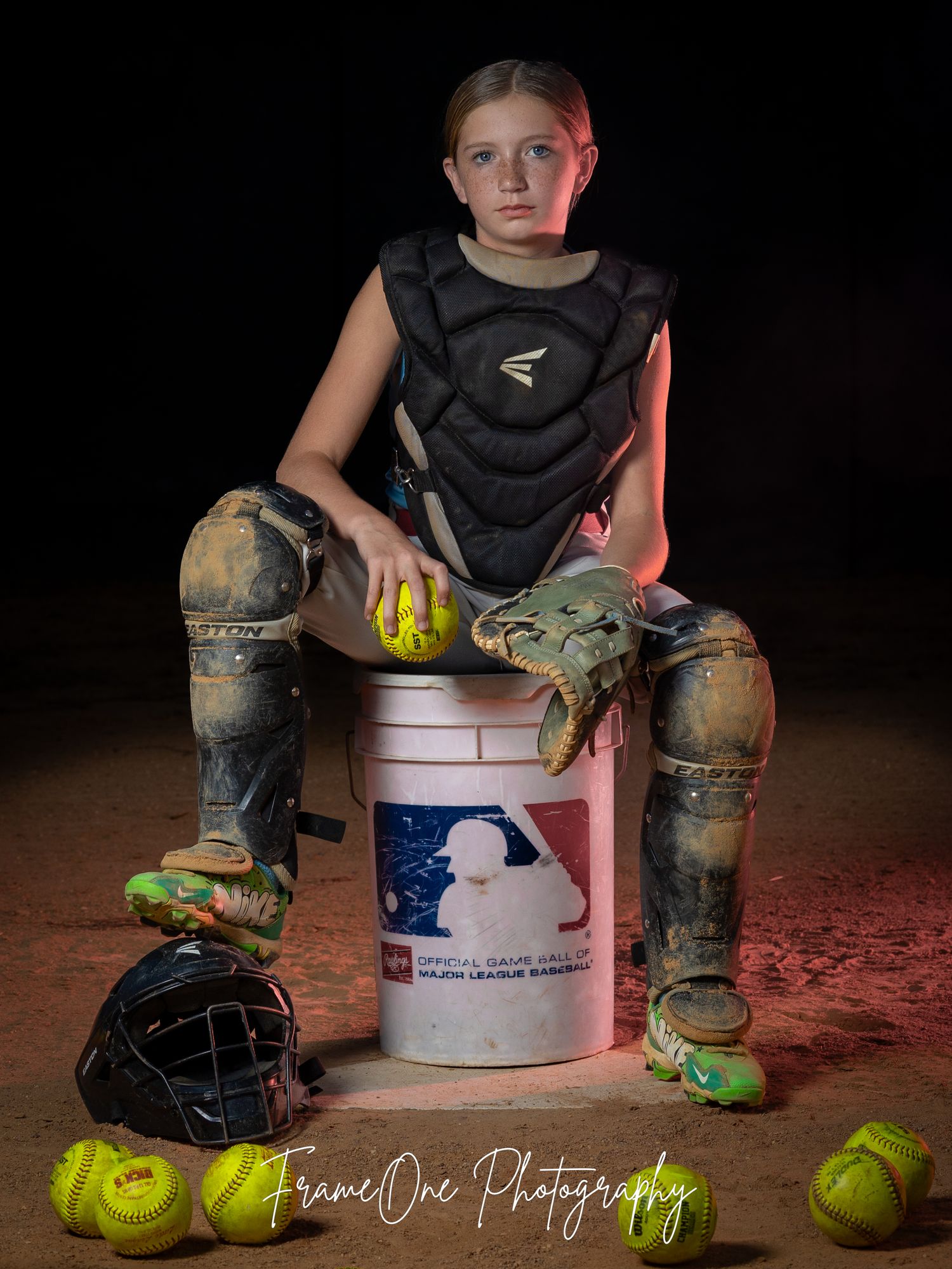 Softball player in protective gear sits on MLB bucket with equipment and balls scattered around in dramatic lighting.