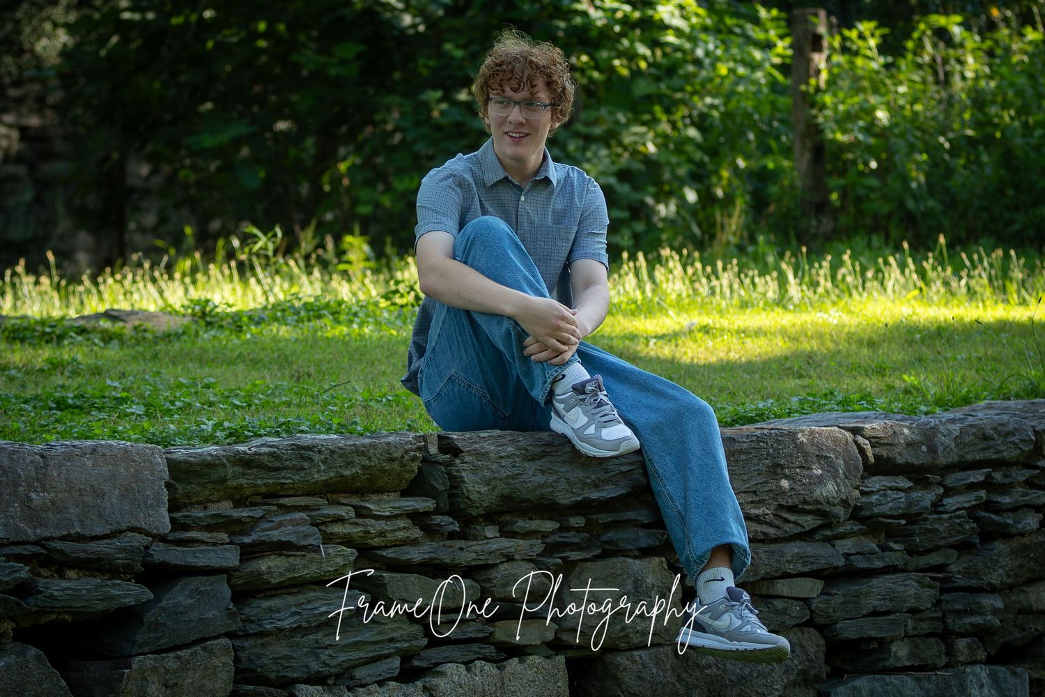 A person in casual blue attire sits casually on a rustic stone wall surrounded by sunlit greenery.