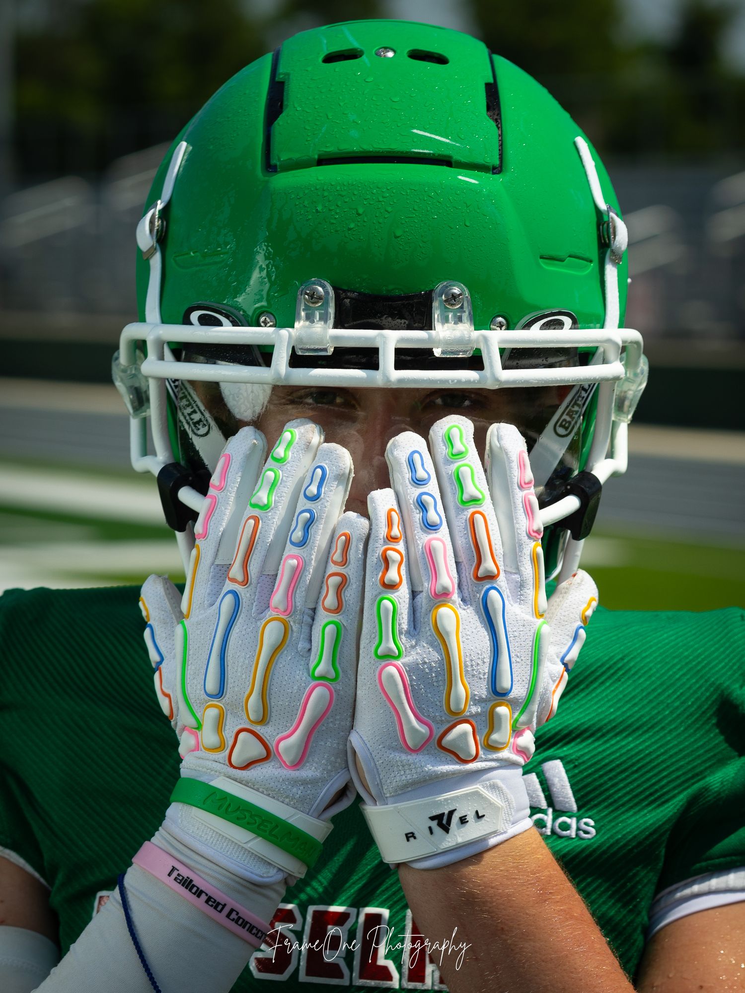 White and pastel colored football gloves with metallic details are displayed on a player wearing a bright green football helmet.