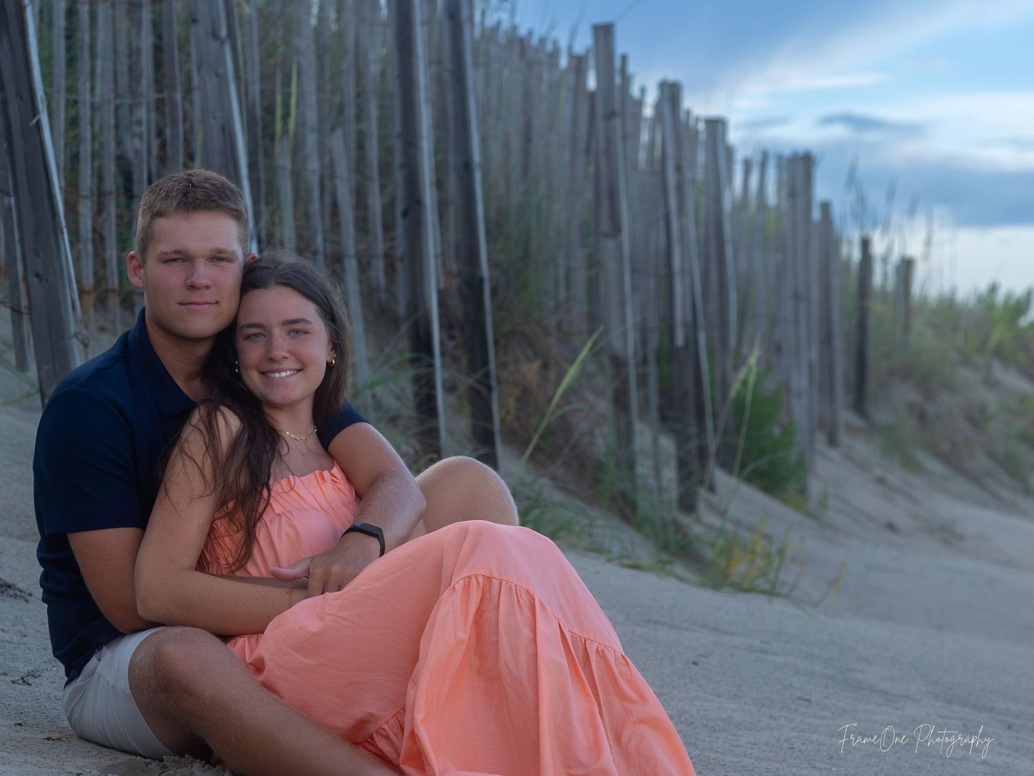 A couple in matching navy and coral outfits sits on a sandy beach near a boardwalk during sunset.