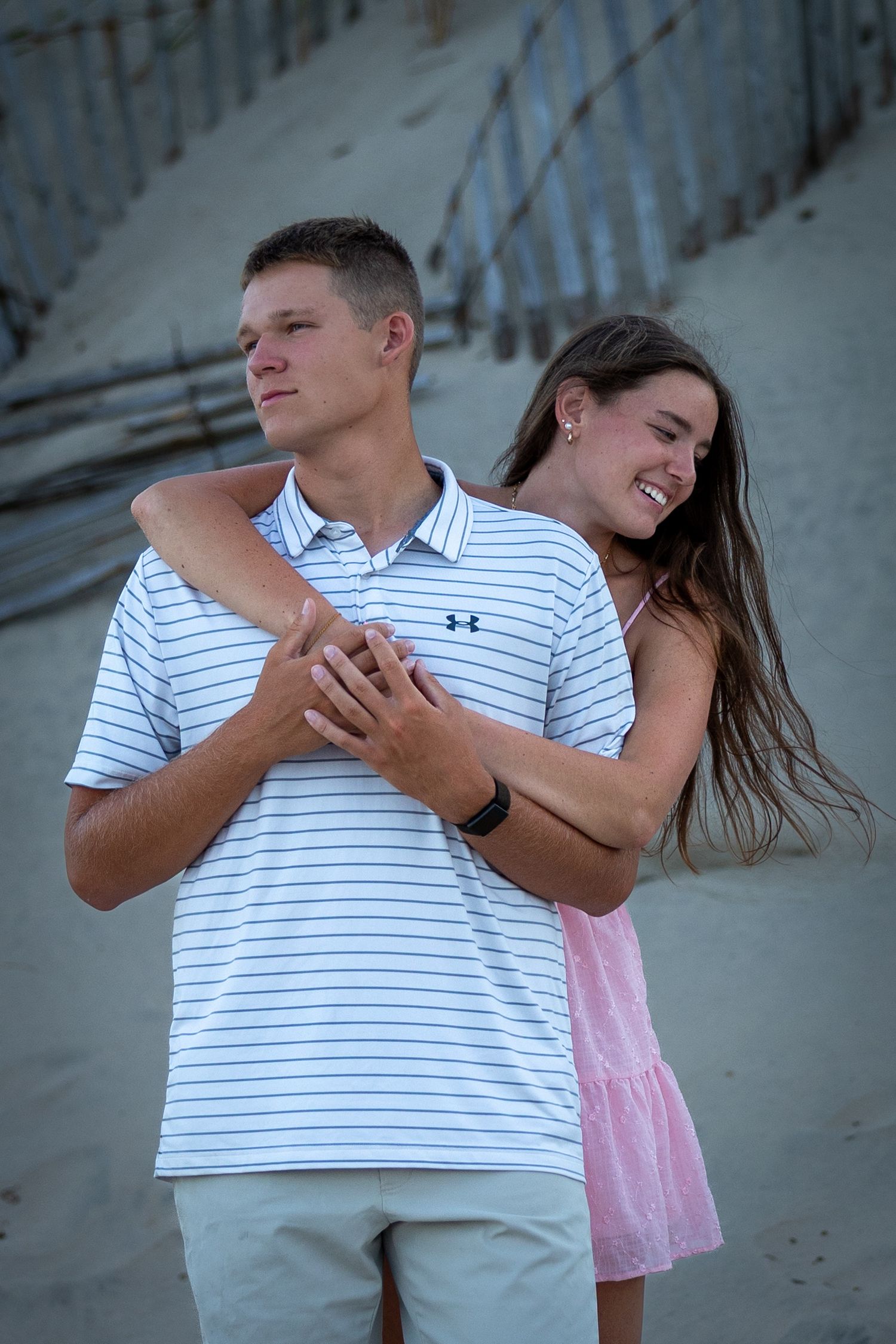 A couple embraces on the beach at dusk, with the person in a pink dress hugging their partner who wears a striped polo shirt.