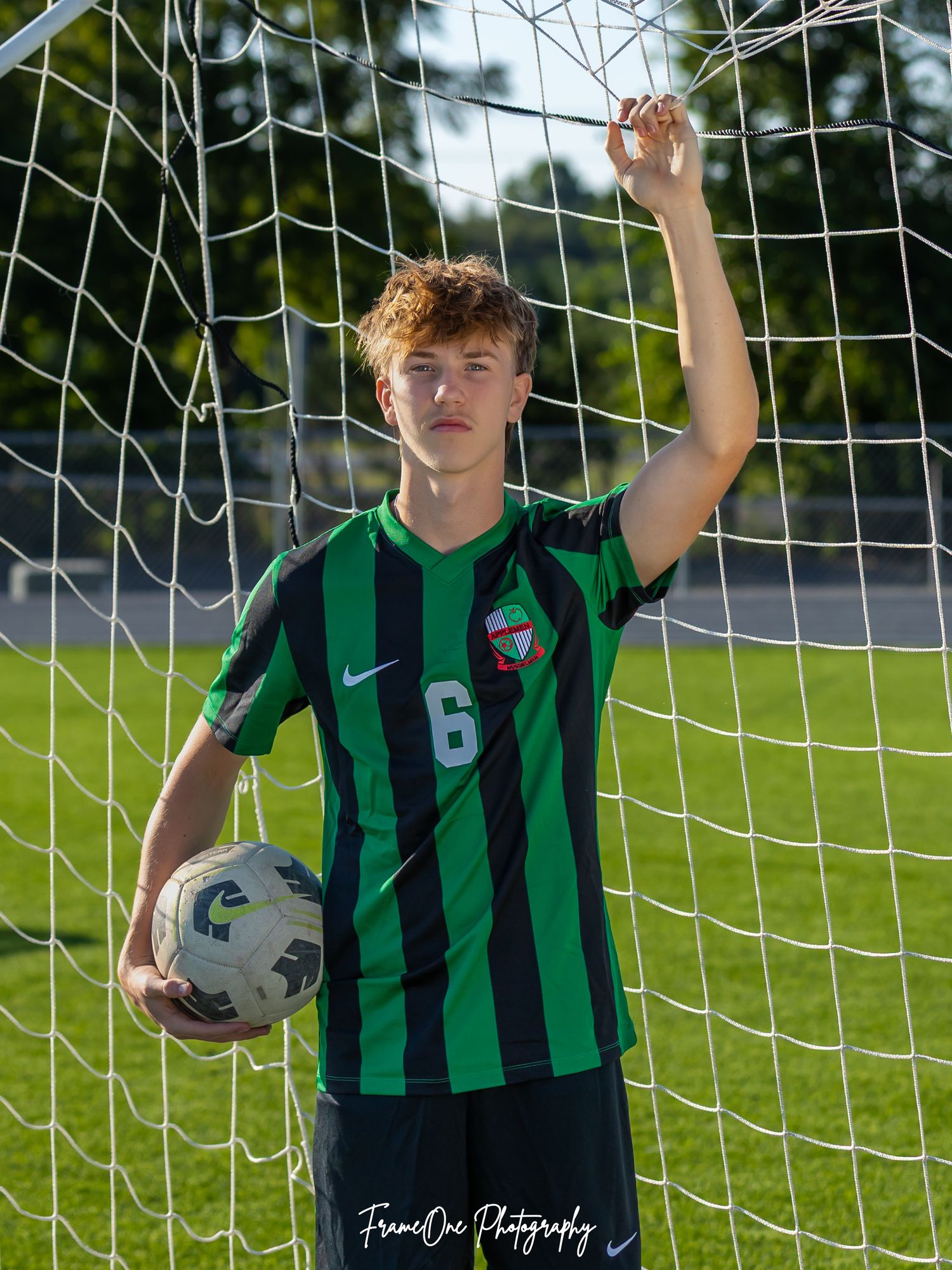 Soccer player in green and black striped uniform #6 poses with soccer ball by goal net during outdoor photo session.