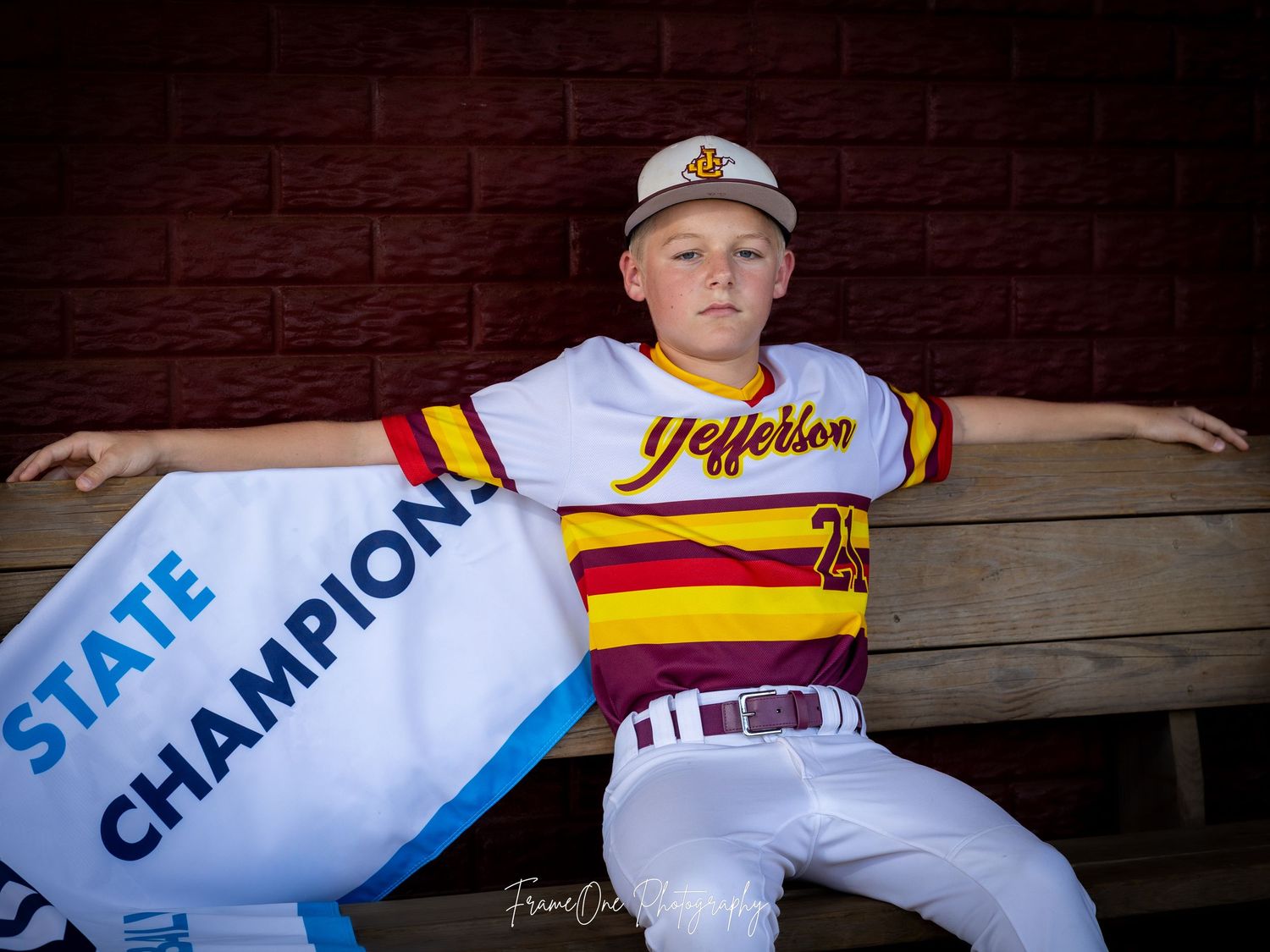 Baseball player in yellow striped jersey and white pants poses in dugout with state champion banner.