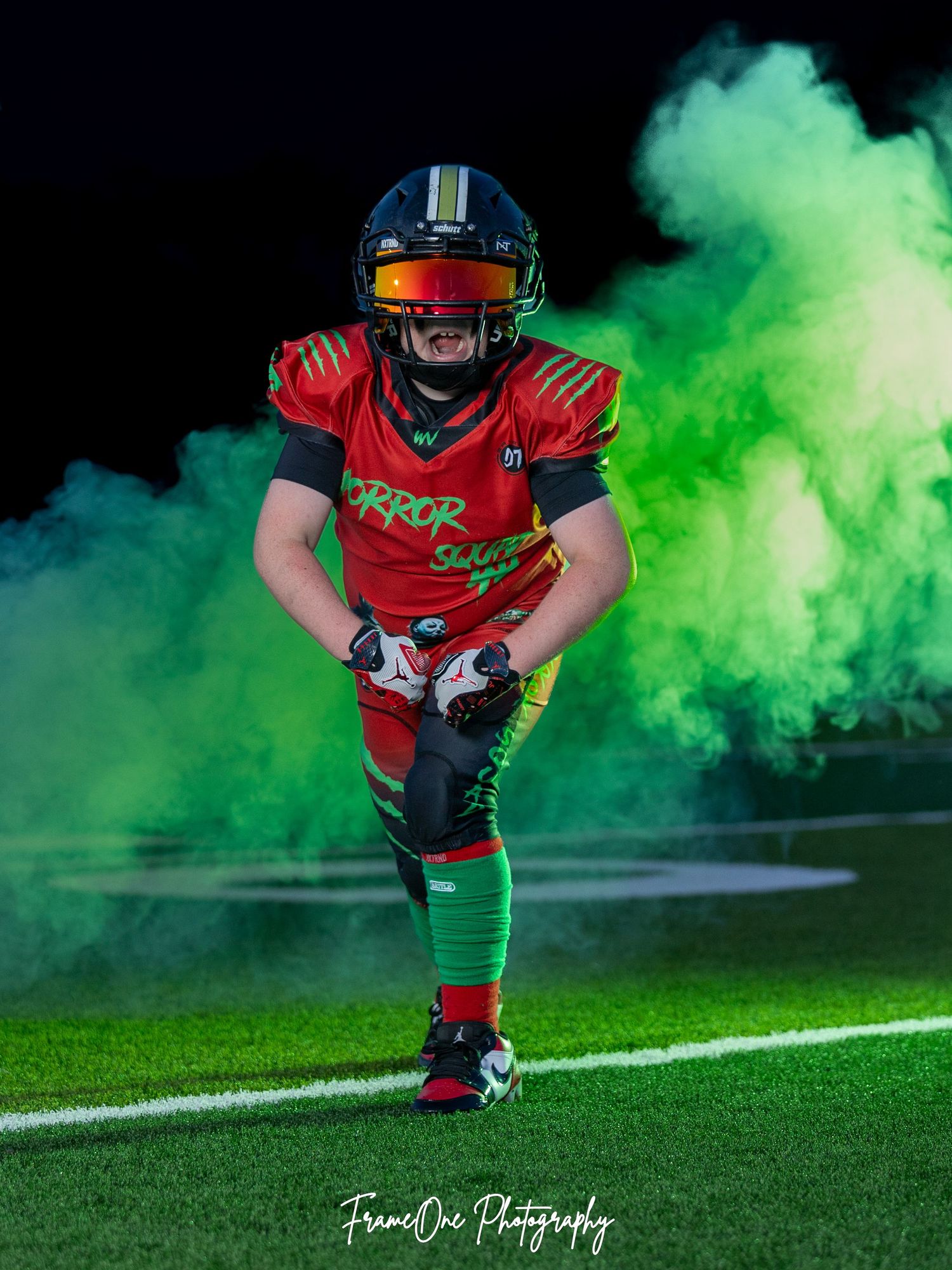 A football player in red uniform poses dramatically in green smoke during a photo shoot on a football field.