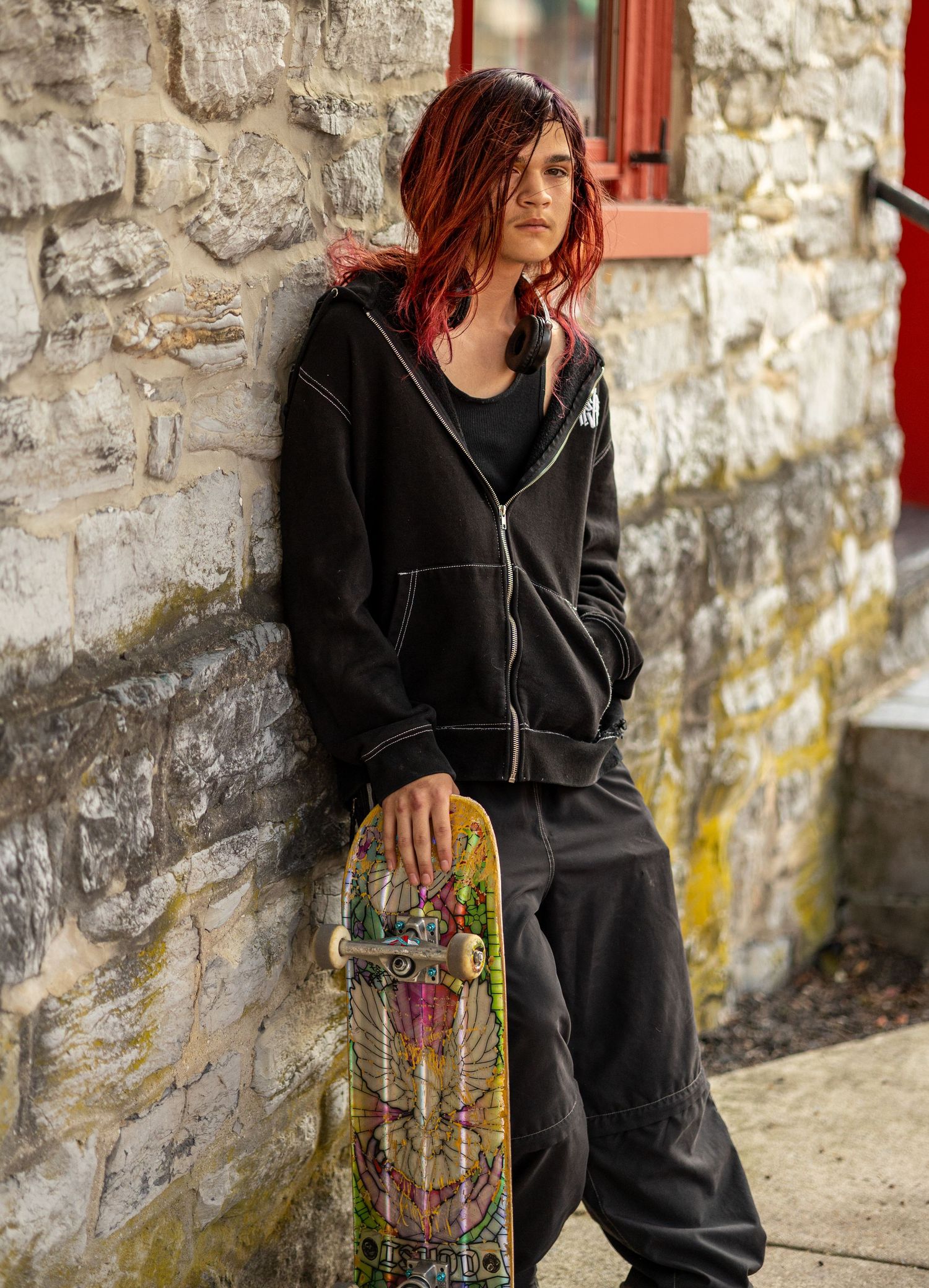 Skater in black hoodie and pants holding a skateboard against stone wall backdrop.
