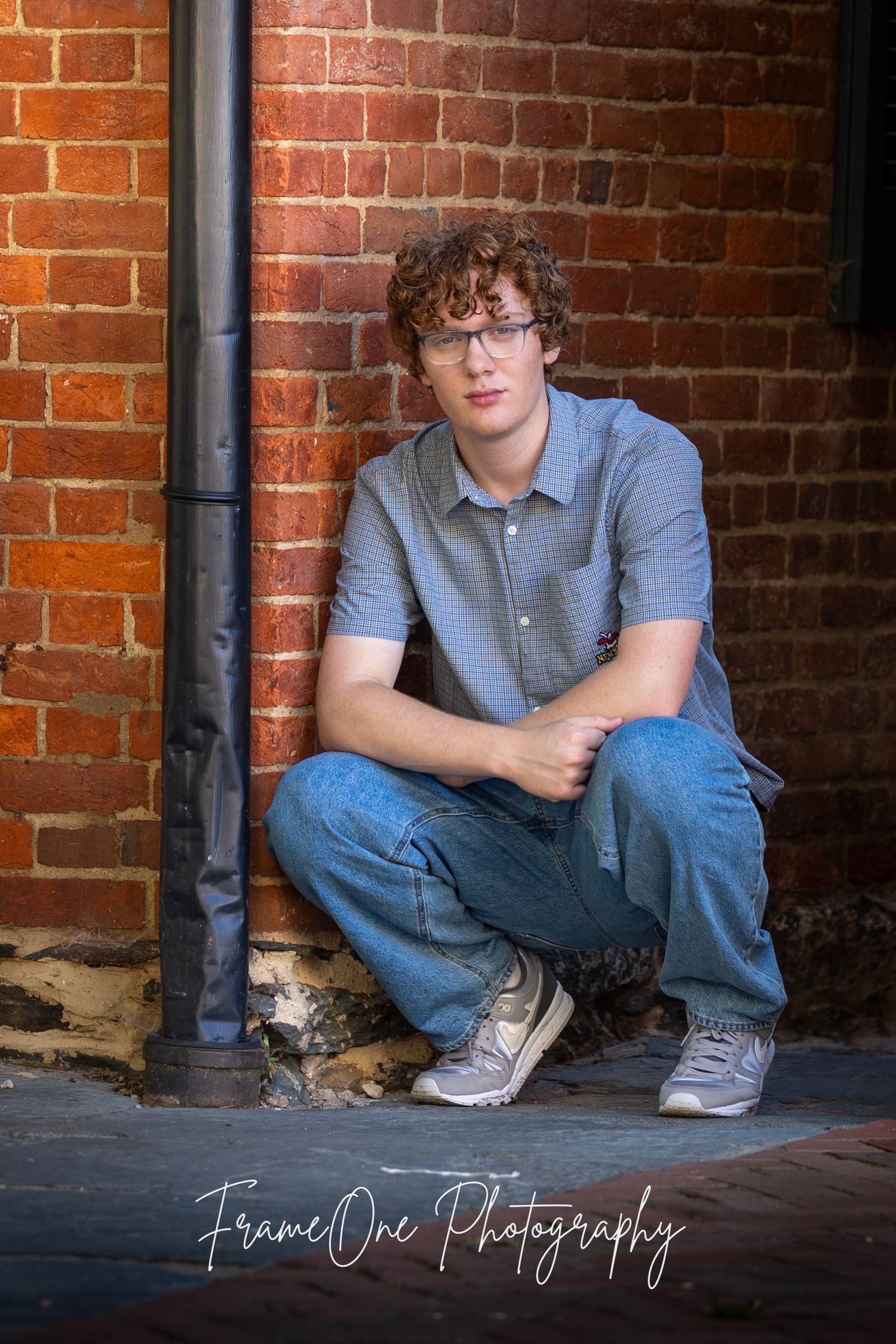 A casual portrait of someone in glasses and a gray polo shirt crouching near a brick wall with dramatic lighting.