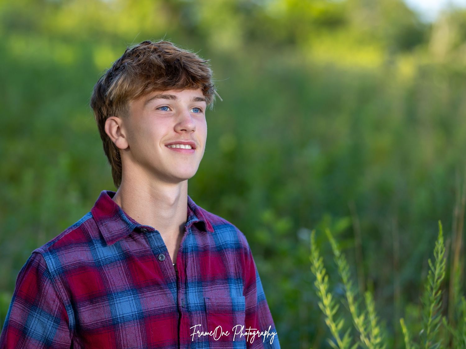 A portrait of a teen in a red and blue plaid flannel shirt smiling outdoors with a soft blurred green background.