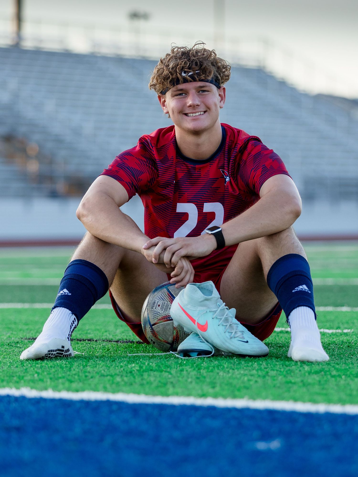 American football player in red jersey number 22 sits on artificial turf field in stadium during sports portrait session.