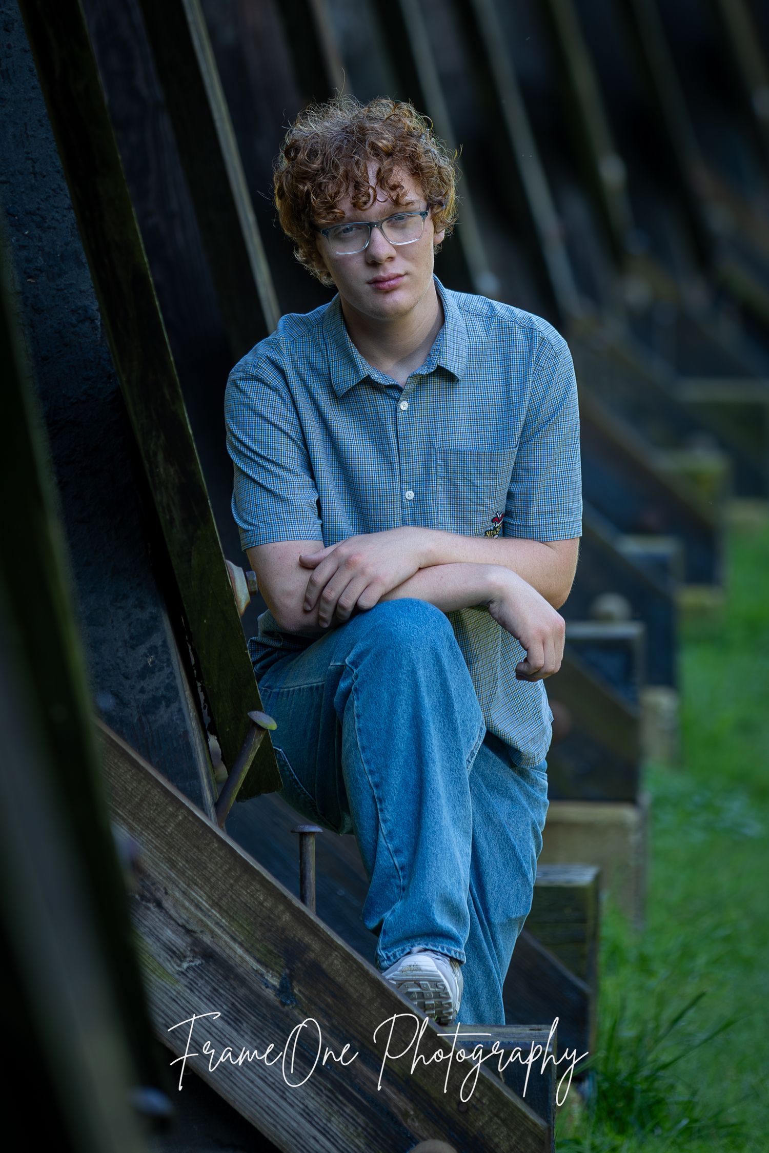 Person in blue plaid shirt and jeans sitting on wooden bench in moody outdoor setting with watermark.