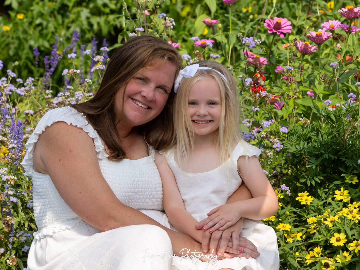 Family members in white outfits sit together among colorful summer flowers and lush greenery in a garden setting.