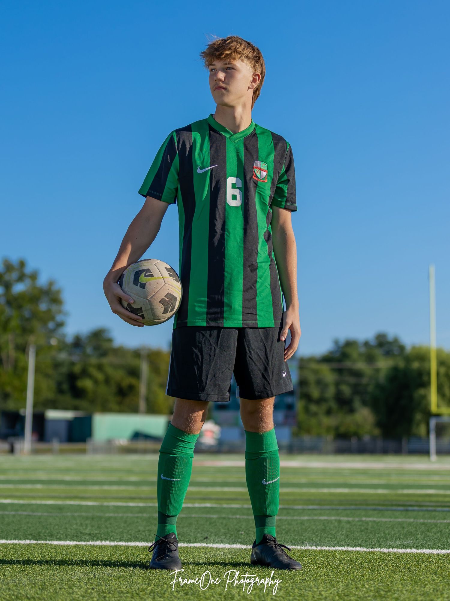 Soccer player in green and black striped jersey with number 6 holds ball while standing on artificial turf field.