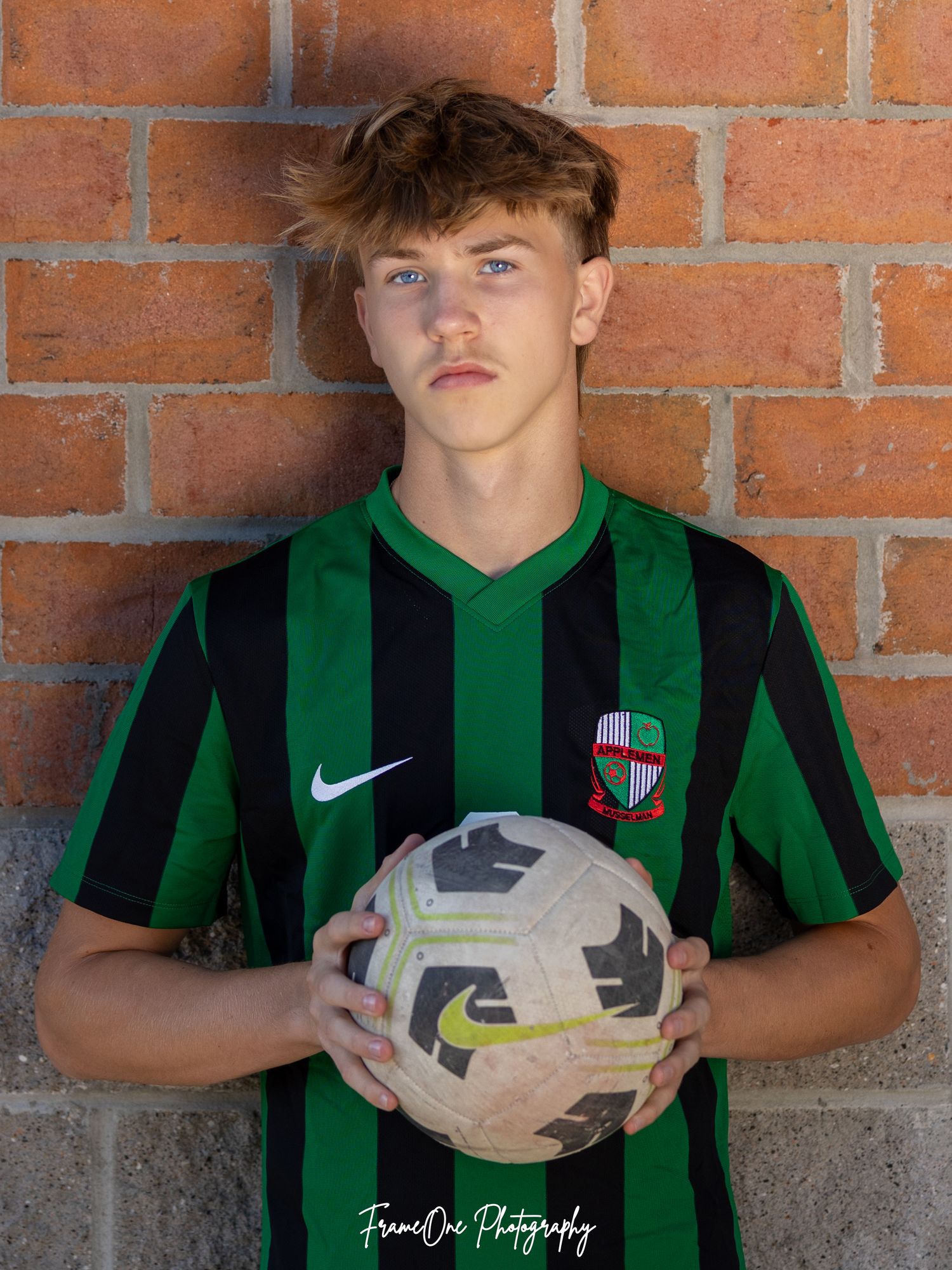 Soccer player in green and black striped Nike jersey poses against brick wall with soccer ball.