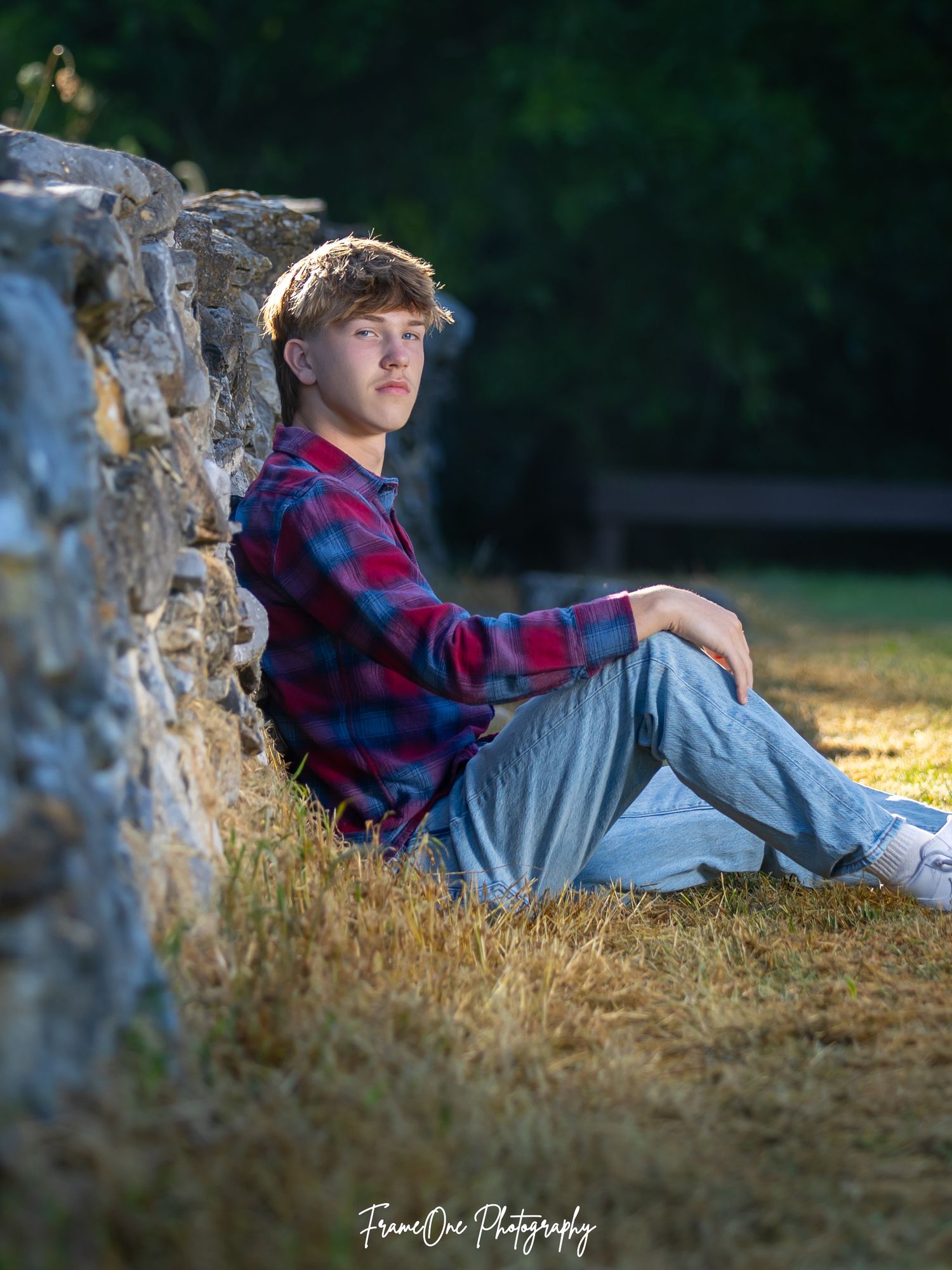 Young person in red plaid flannel shirt and jeans reclines against a stone wall at sunset in an outdoor portrait setting.