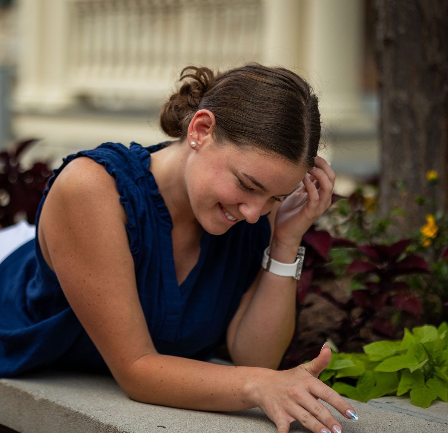 Someone in a navy blue sleeveless top laughs while looking down at the ground near some greenery and flowers.