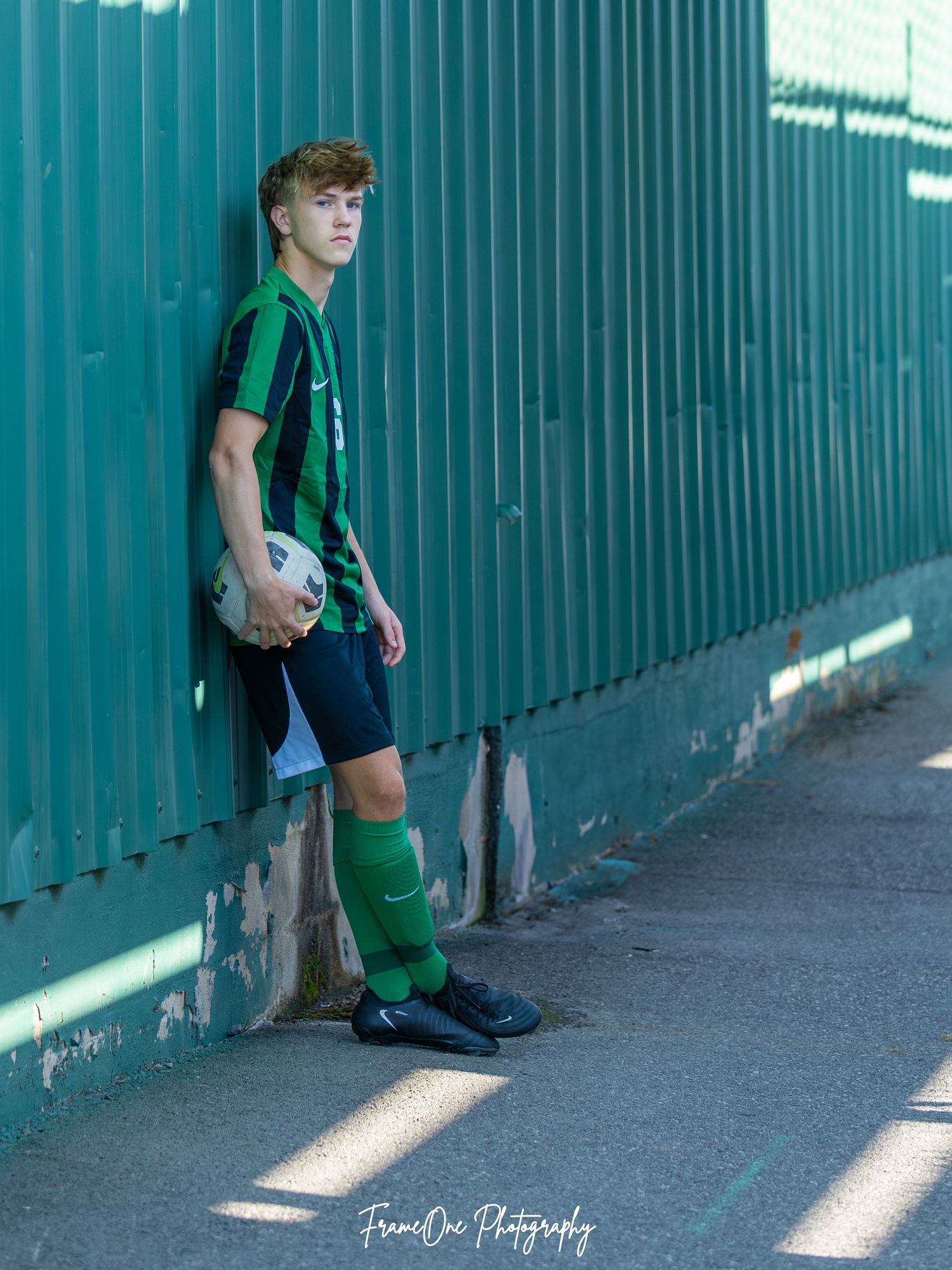Soccer player in green and black uniform leans against teal fence in dramatic lighting.