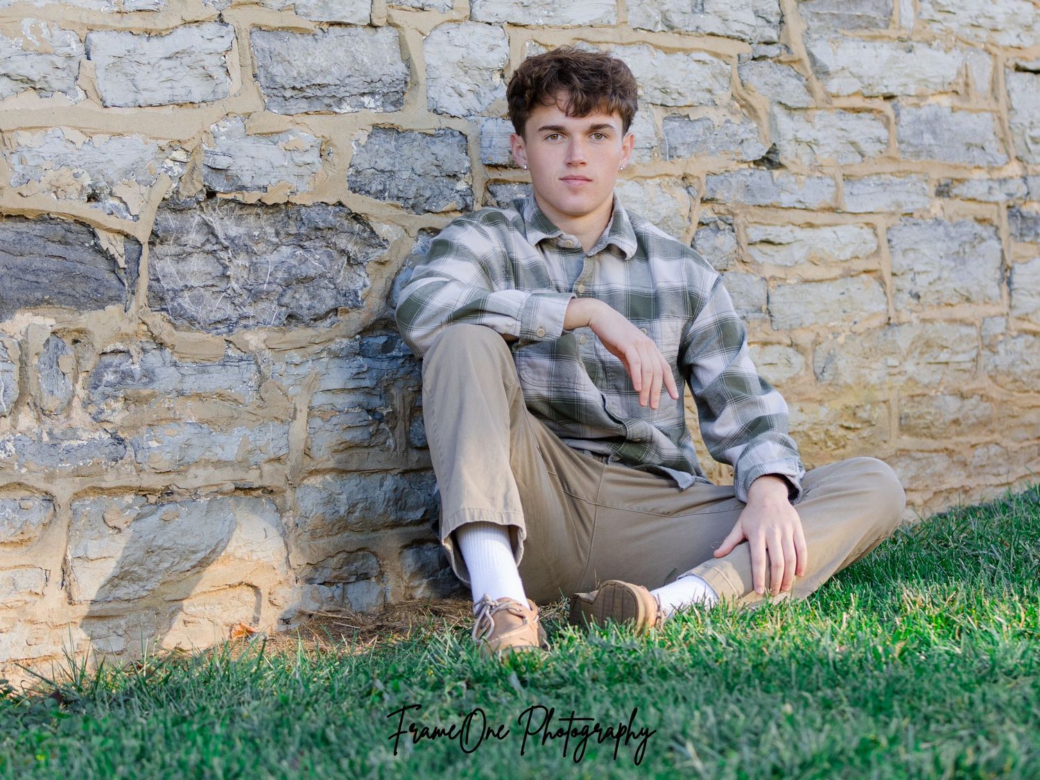 Person in plaid shirt and khakis lounges against stone wall on grassy slope during outdoor photo session.
