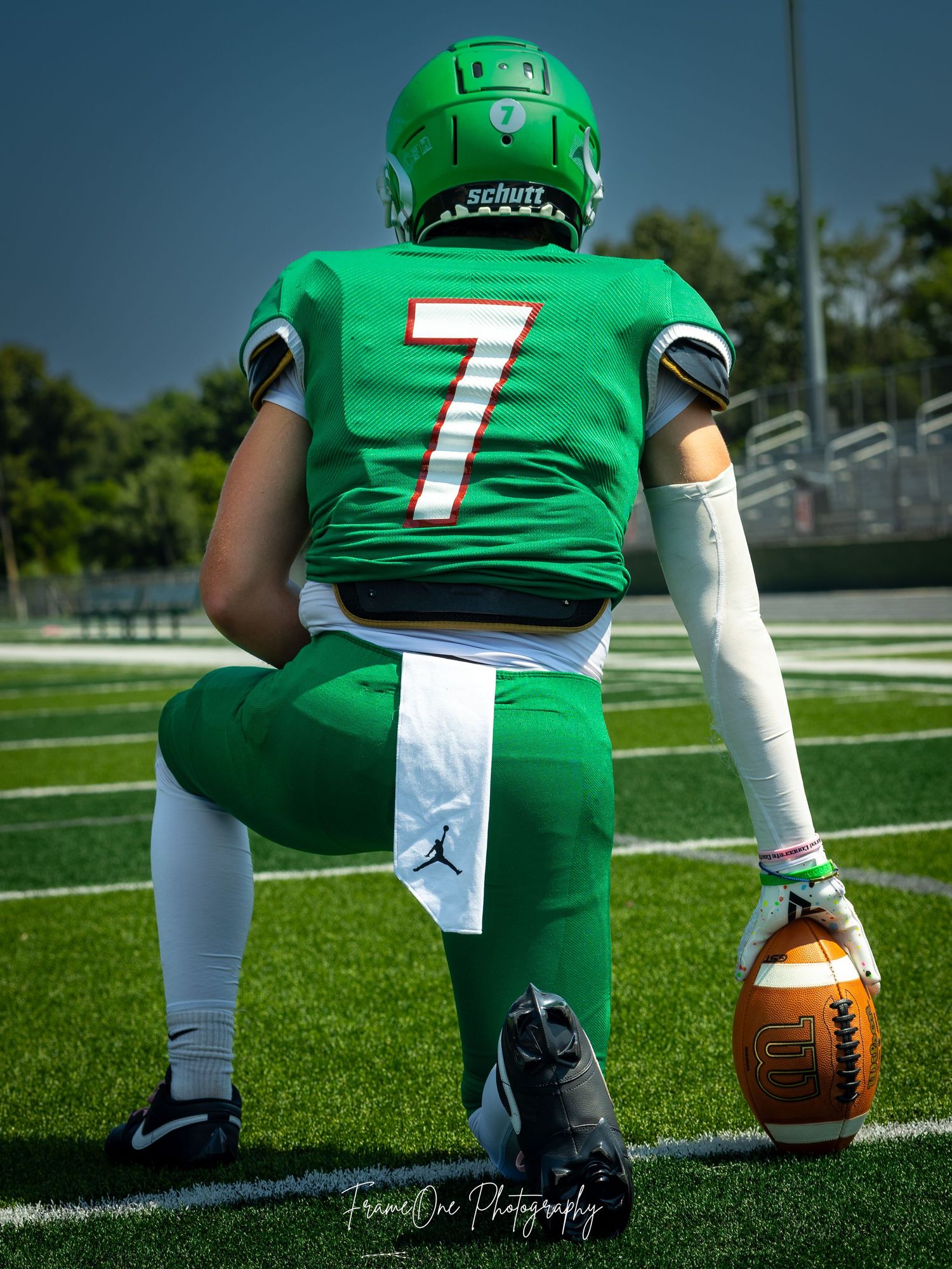 Back view of football player wearing green jersey number 7 kneeling on artificial turf with ball.
