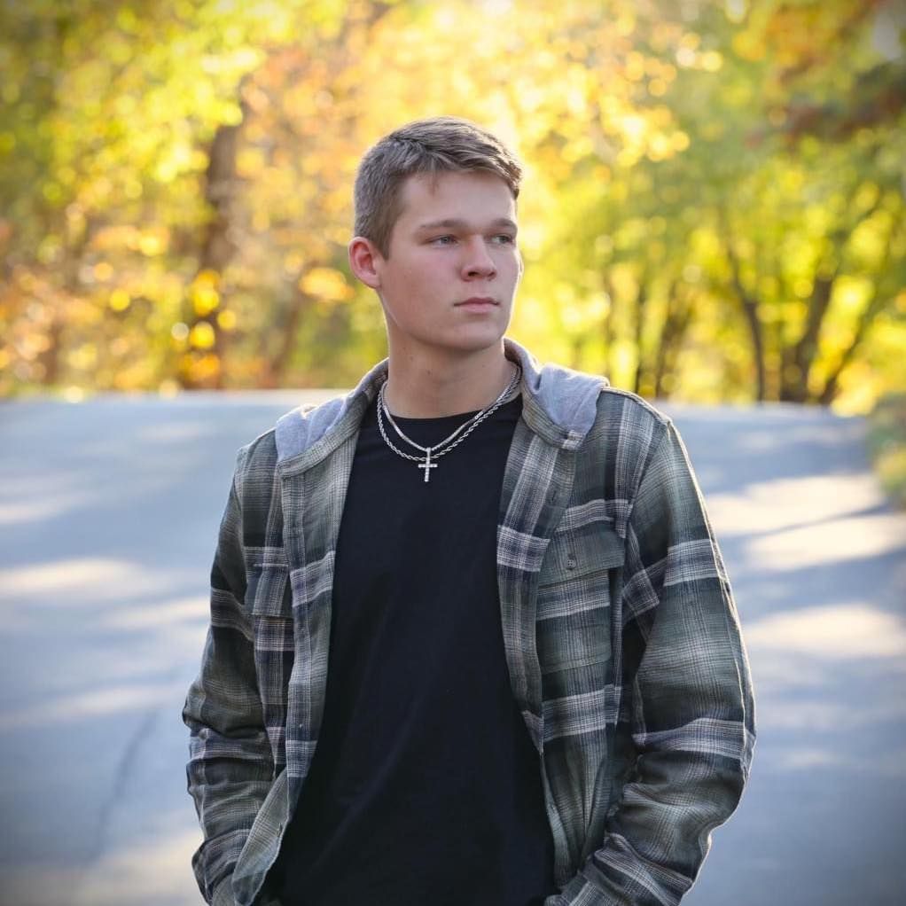 Person wearing plaid jacket stands in autumn forest setting with golden foliage in background during daytime.