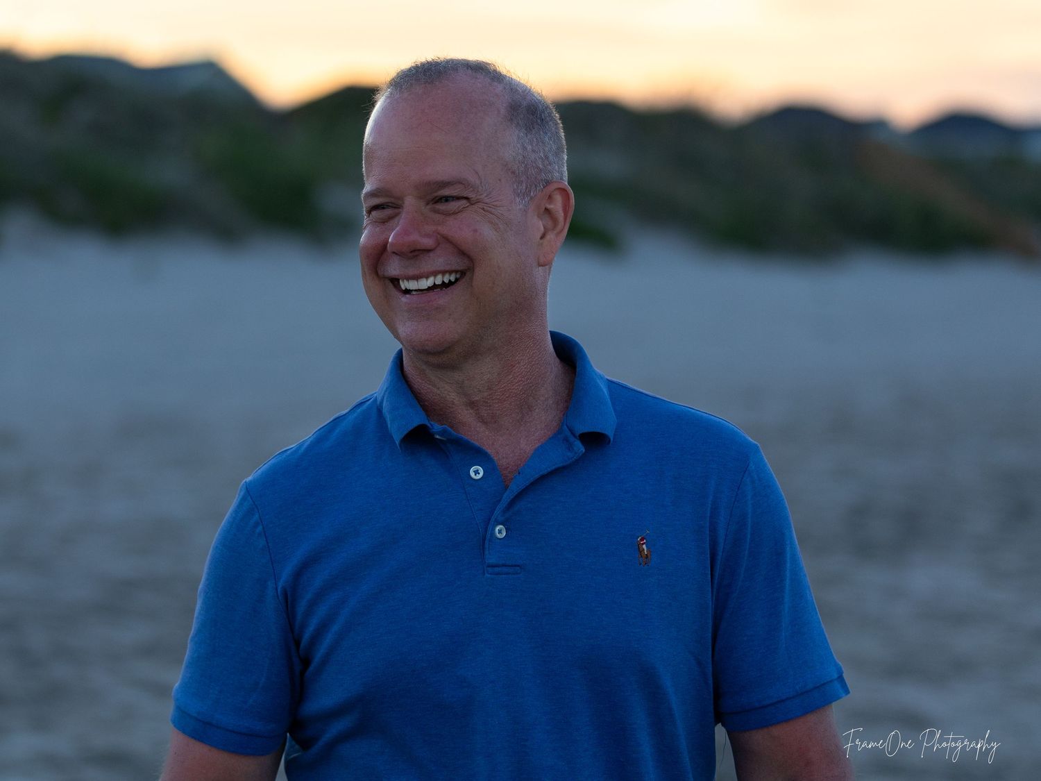Person wearing a blue polo shirt smiles warmly outdoors at sunset with blurred natural background.
