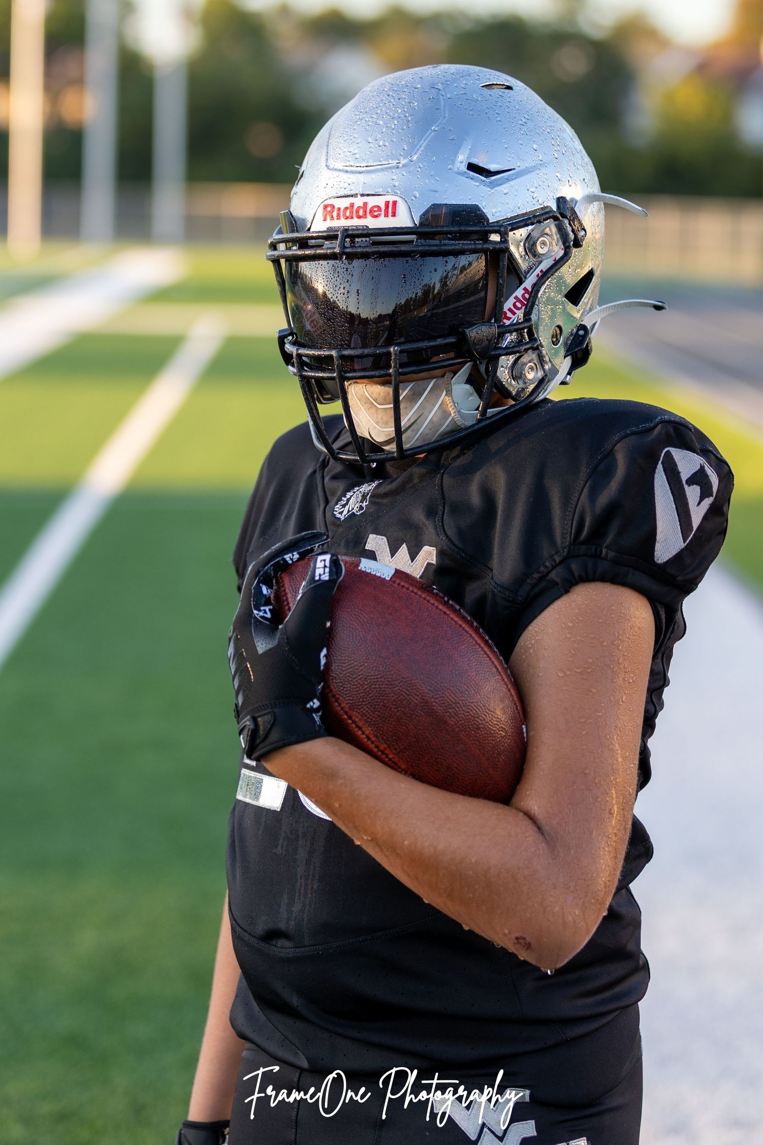 Football player in black uniform carries ball during sunset practice on outdoor field.