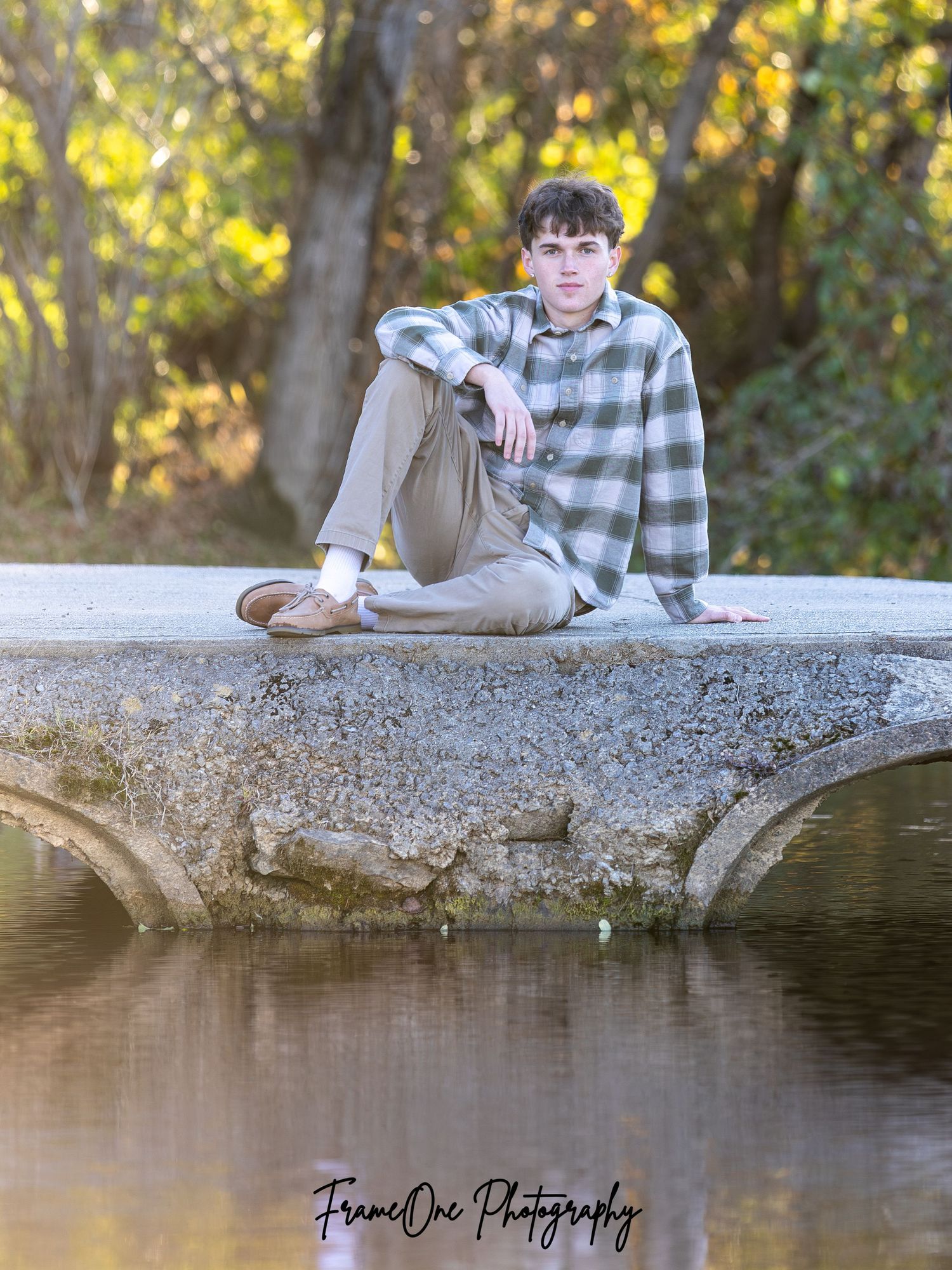 Person rests on concrete ledge near pond during autumn outdoor portrait session with golden foliage in background.
