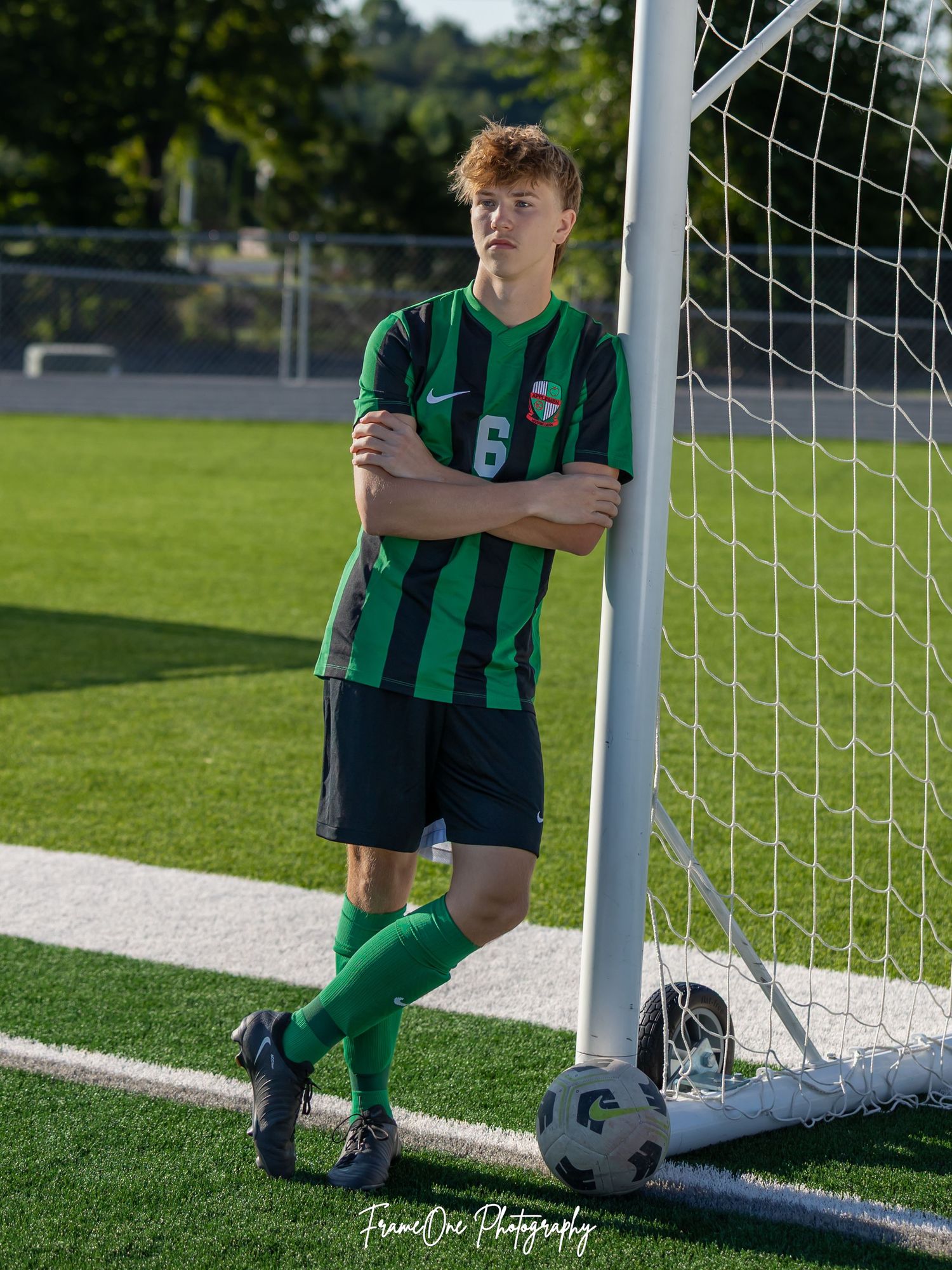 Soccer player in green striped uniform poses by goal post on sunny day.