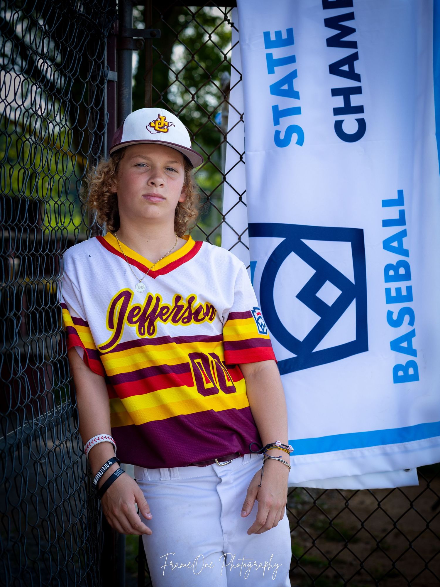 Baseball player in Jefferson team uniform with white and maroon striped jersey poses next to State Championship banner.