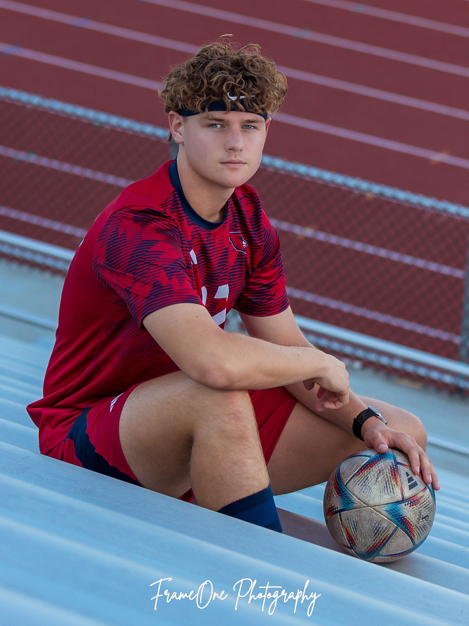 An athlete in a red uniform sits on stadium bleachers holding a soccer ball against a running track background.