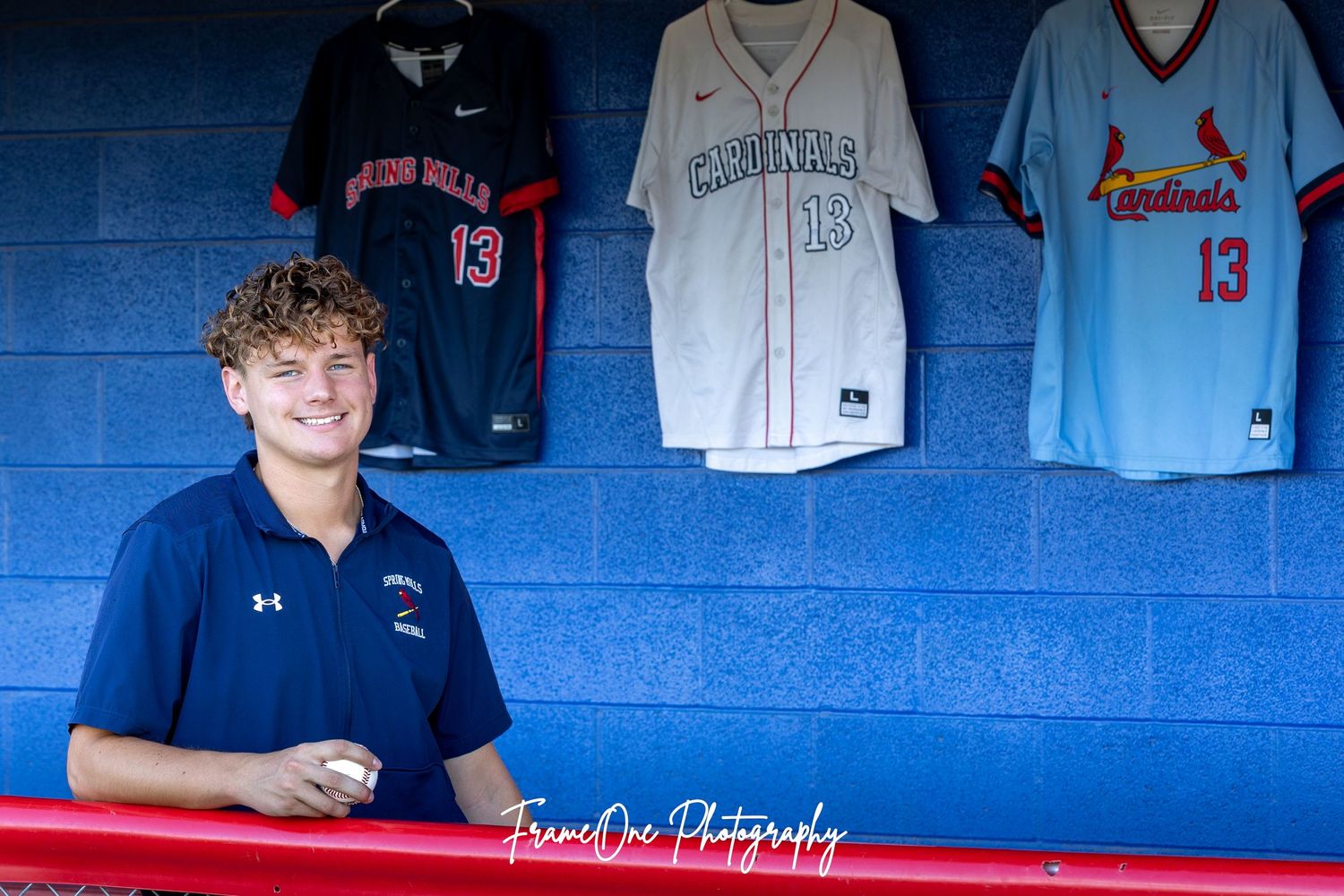 Baseball player stands in dugout with jersey collection displayed on blue wall behind him.