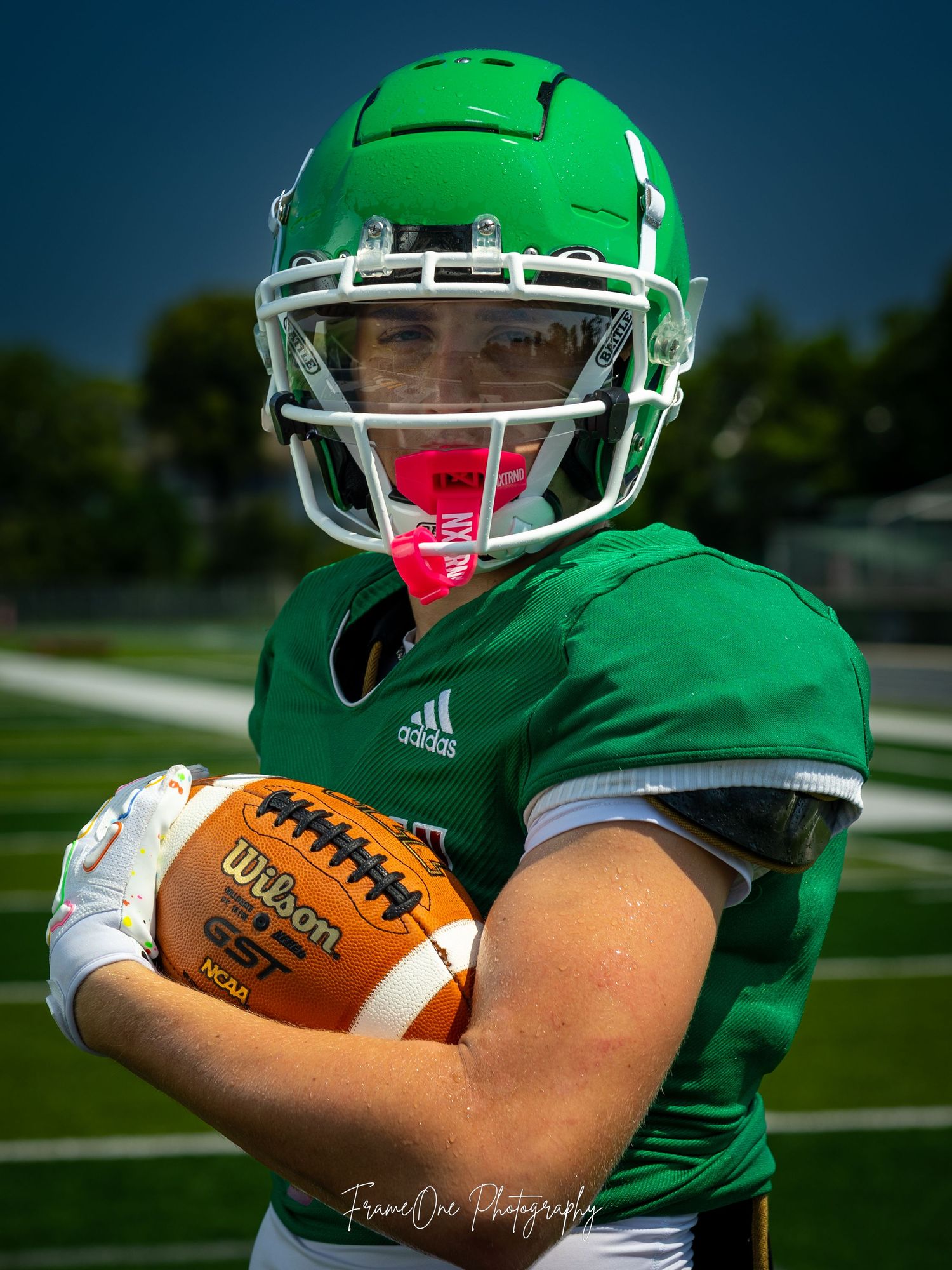 Football player wearing green Adidas jersey and helmet holds Wilson football during outdoor practice session.