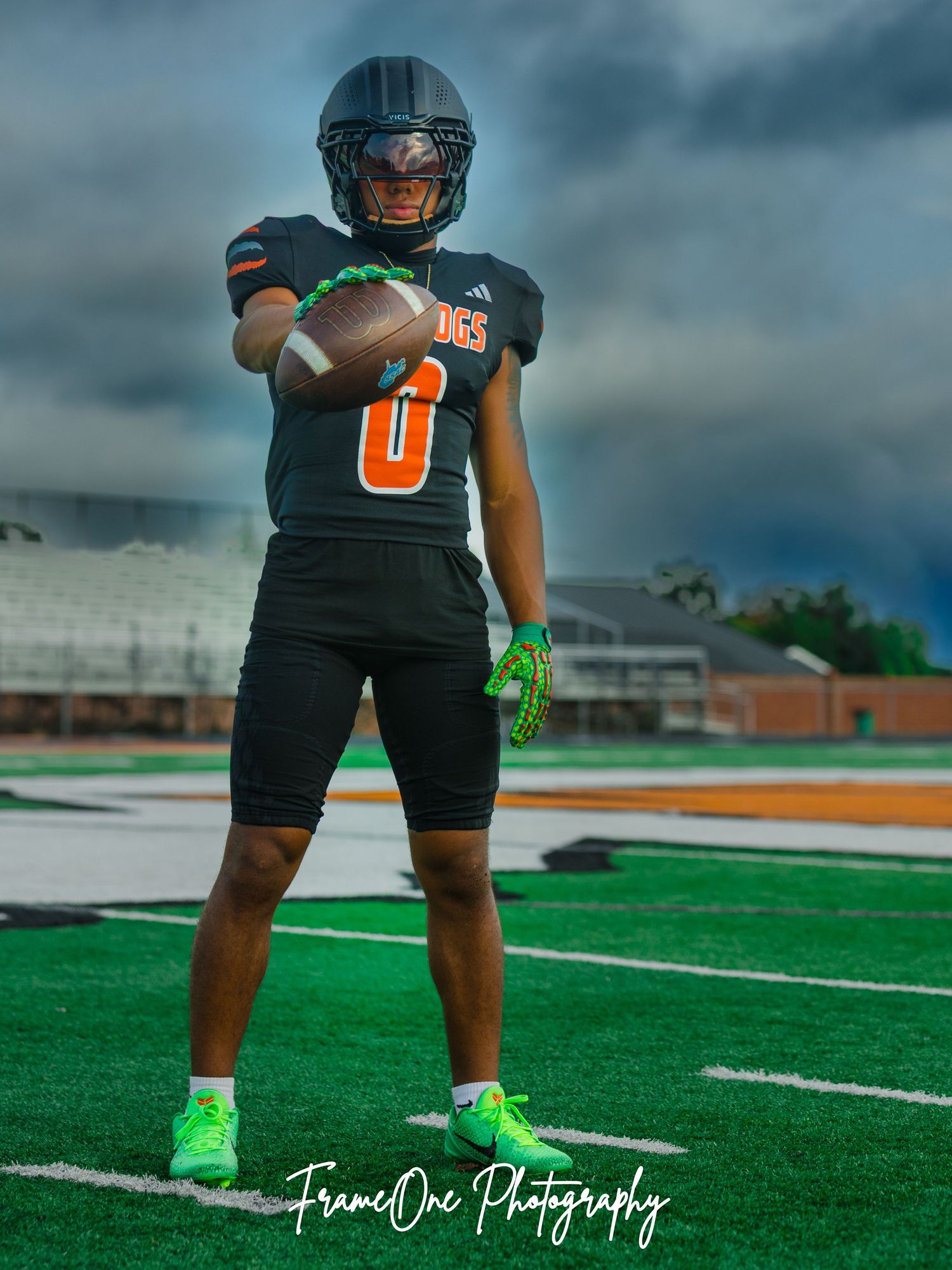 Football player in black uniform and green cleats stands on artificial turf field under dramatic stormy sky.
