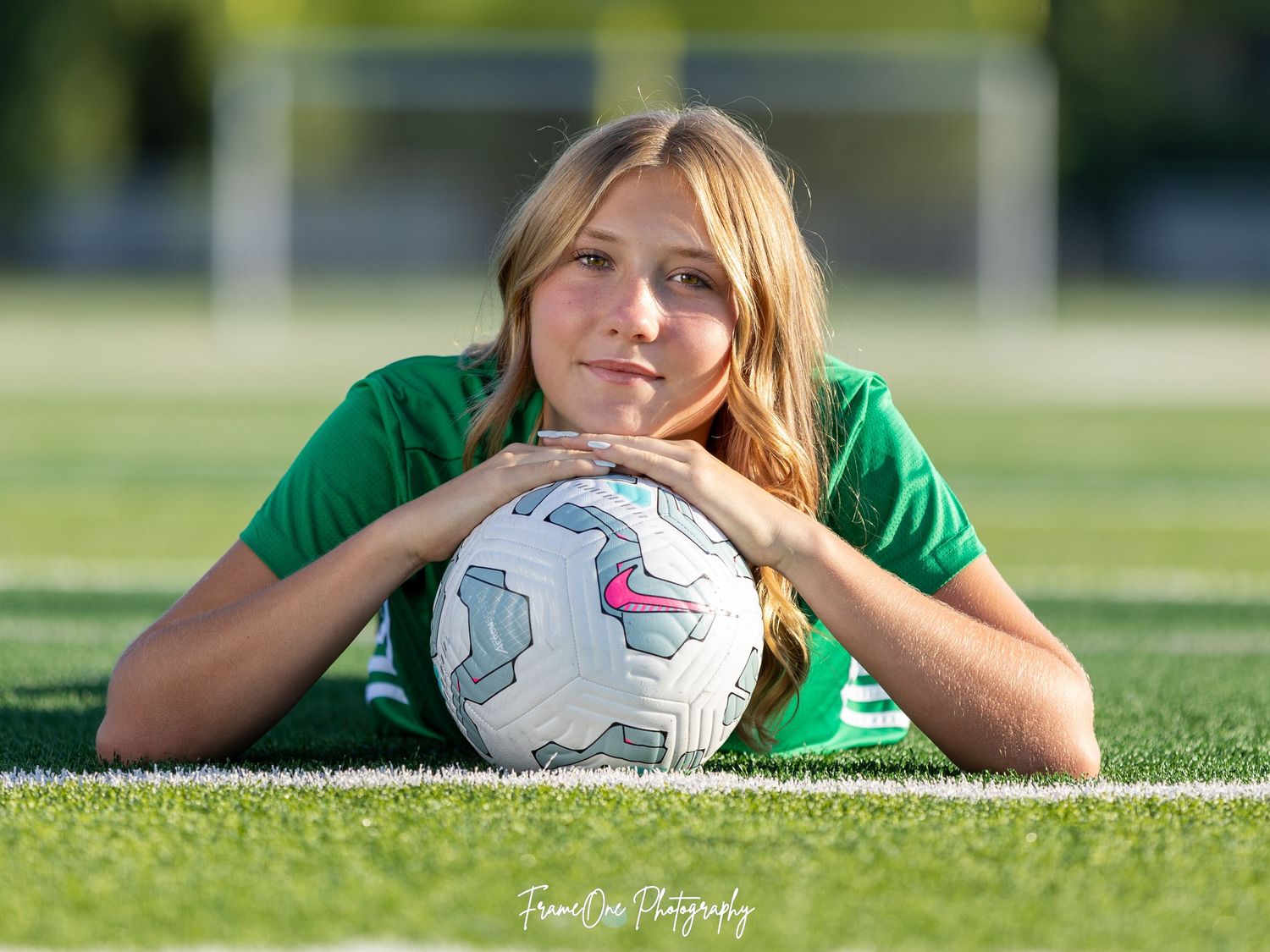Soccer player in green jersey rests on grass field with white soccer ball on sunny day.