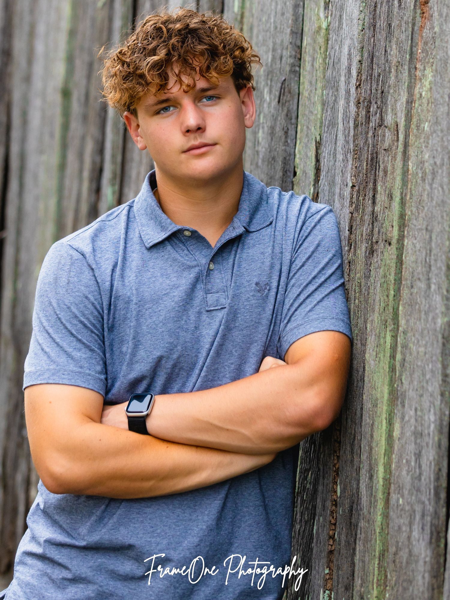 Young person in blue polo shirt poses against rustic wooden fence in casual senior portrait style.