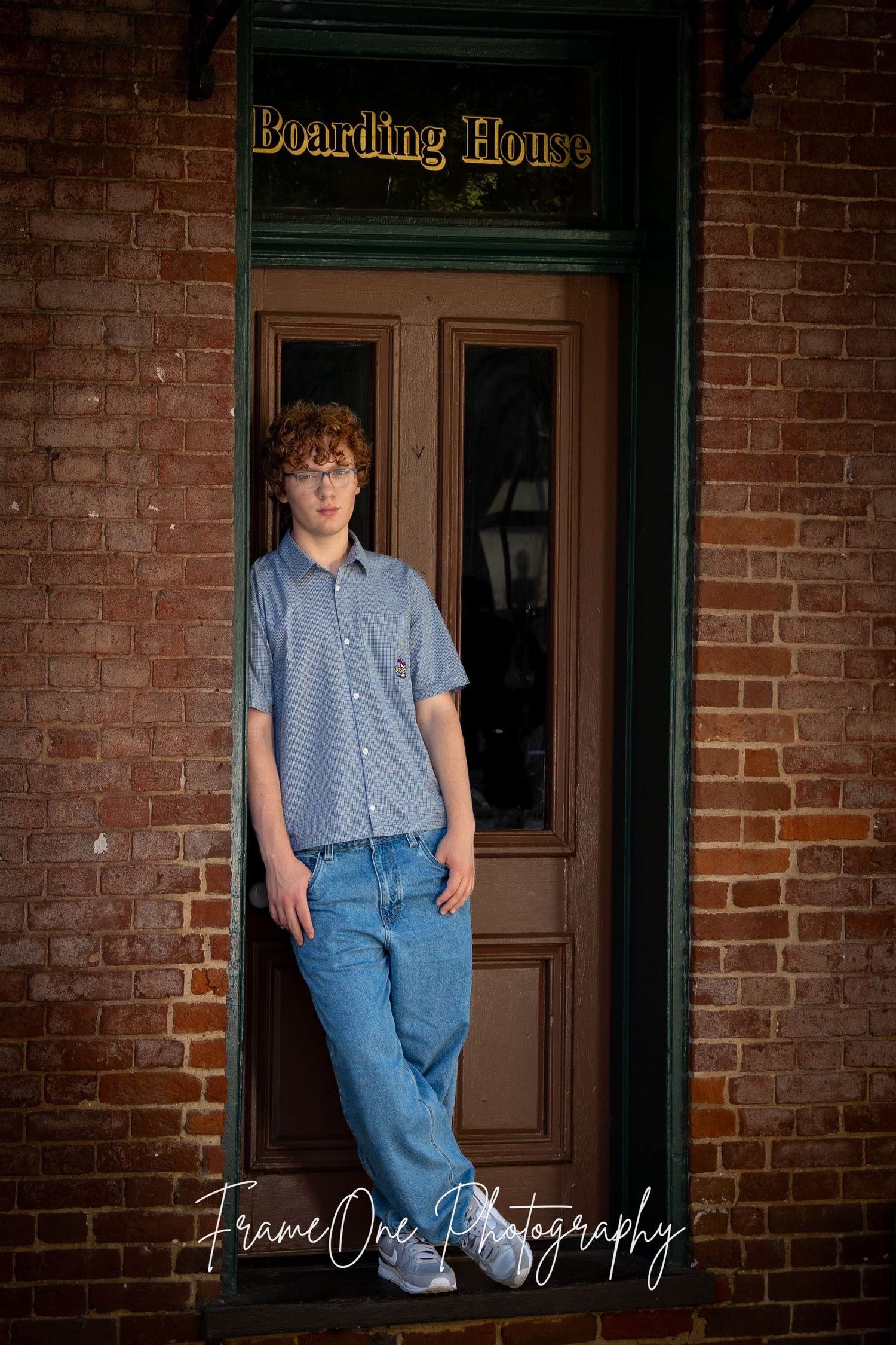 Young person in denim and blue shirt leans against brick wall near vintage boarding house entrance.