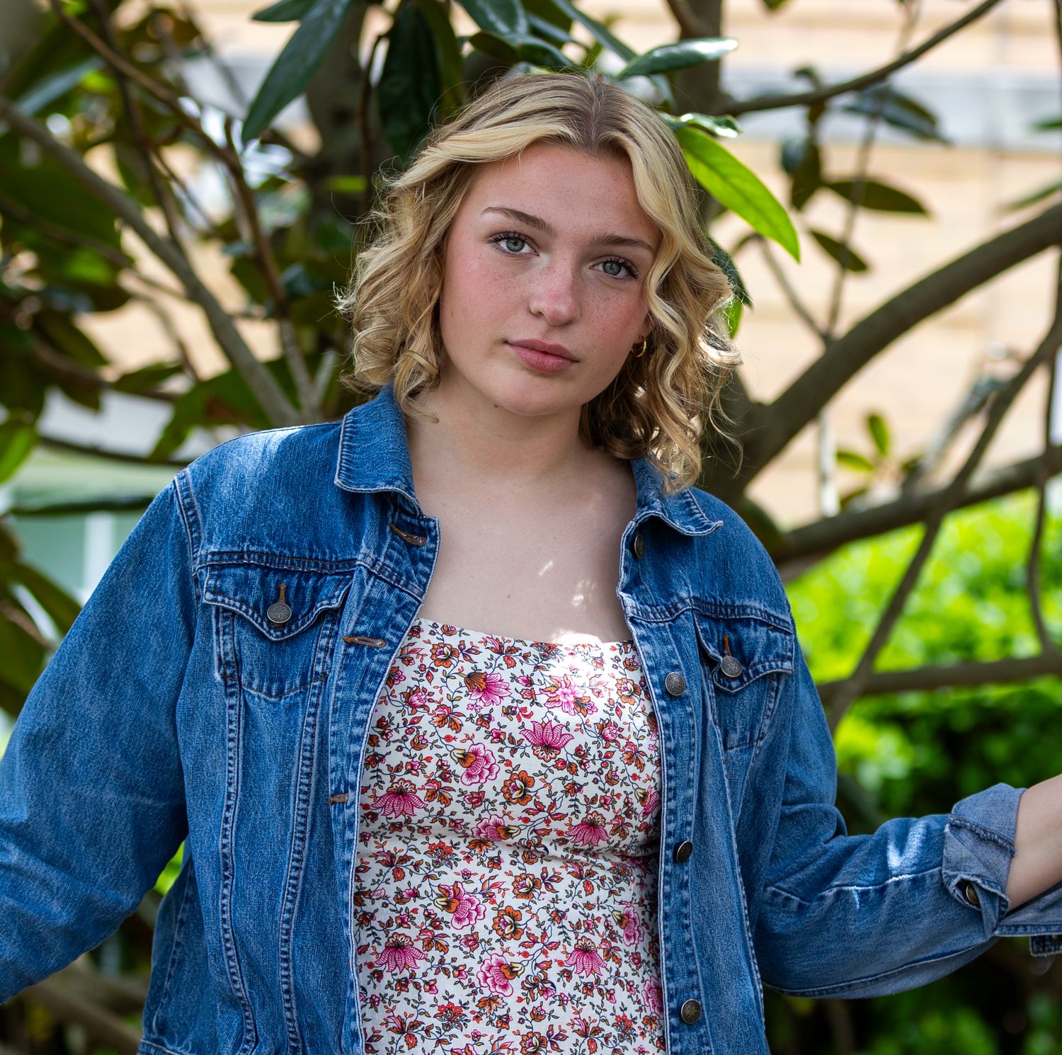 Portrait of a person wearing a denim jacket and floral dress against green foliage.