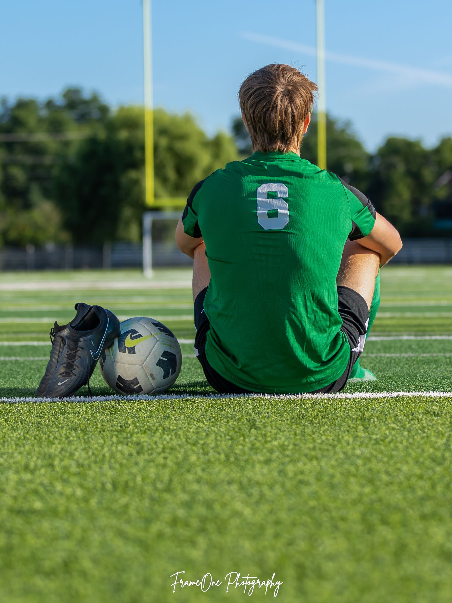 Soccer player wearing number 6 green jersey sits on field with ball and cleats.