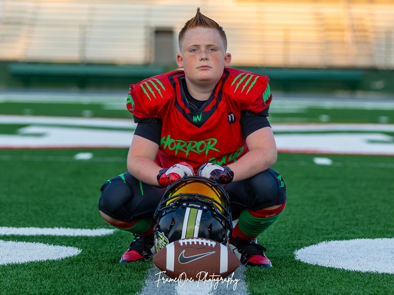 Football player in black and red uniform kneels on field with orange sky and stadium lights in background.