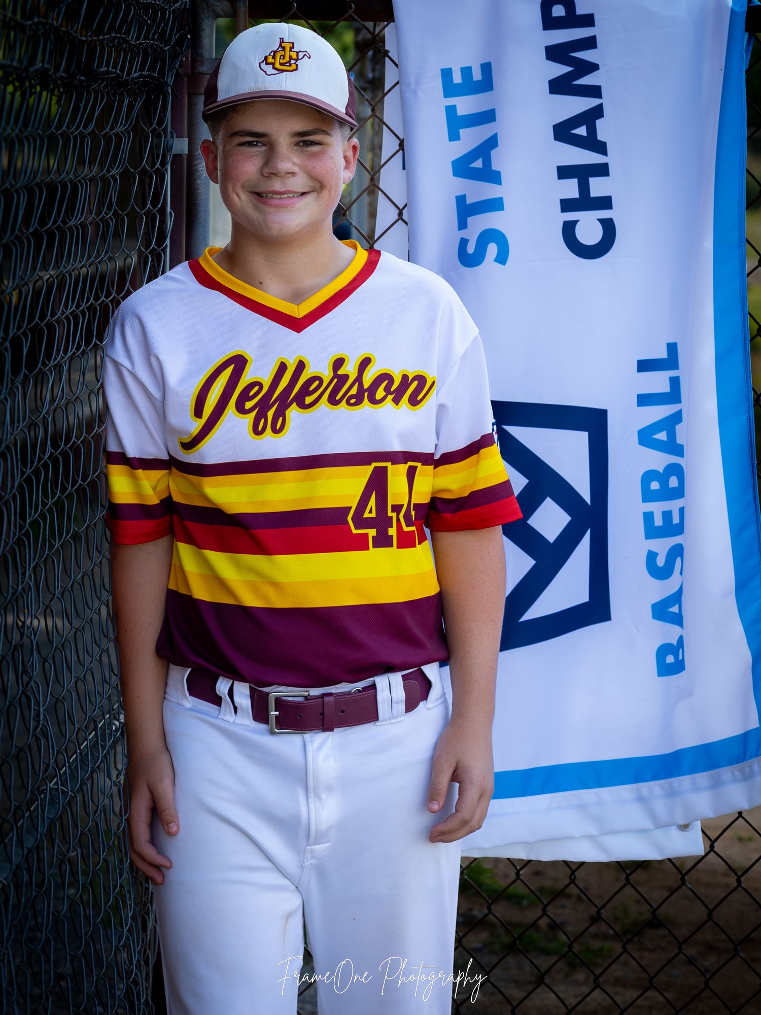 Baseball player in white Jefferson uniform number 44 with burgundy and yellow stripes poses next to championship banner.