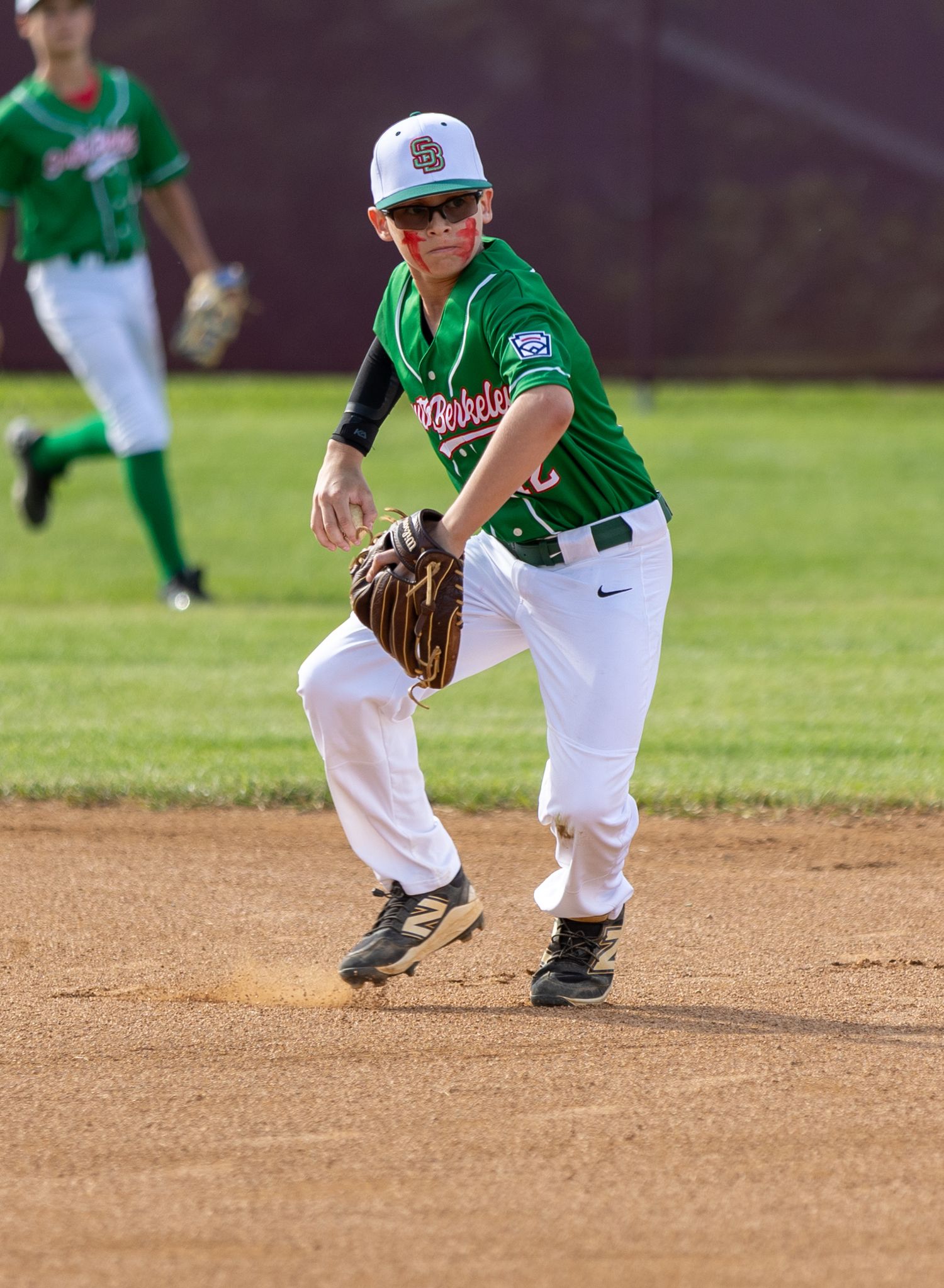 Youth baseball player in green uniform fielding position on dirt infield.