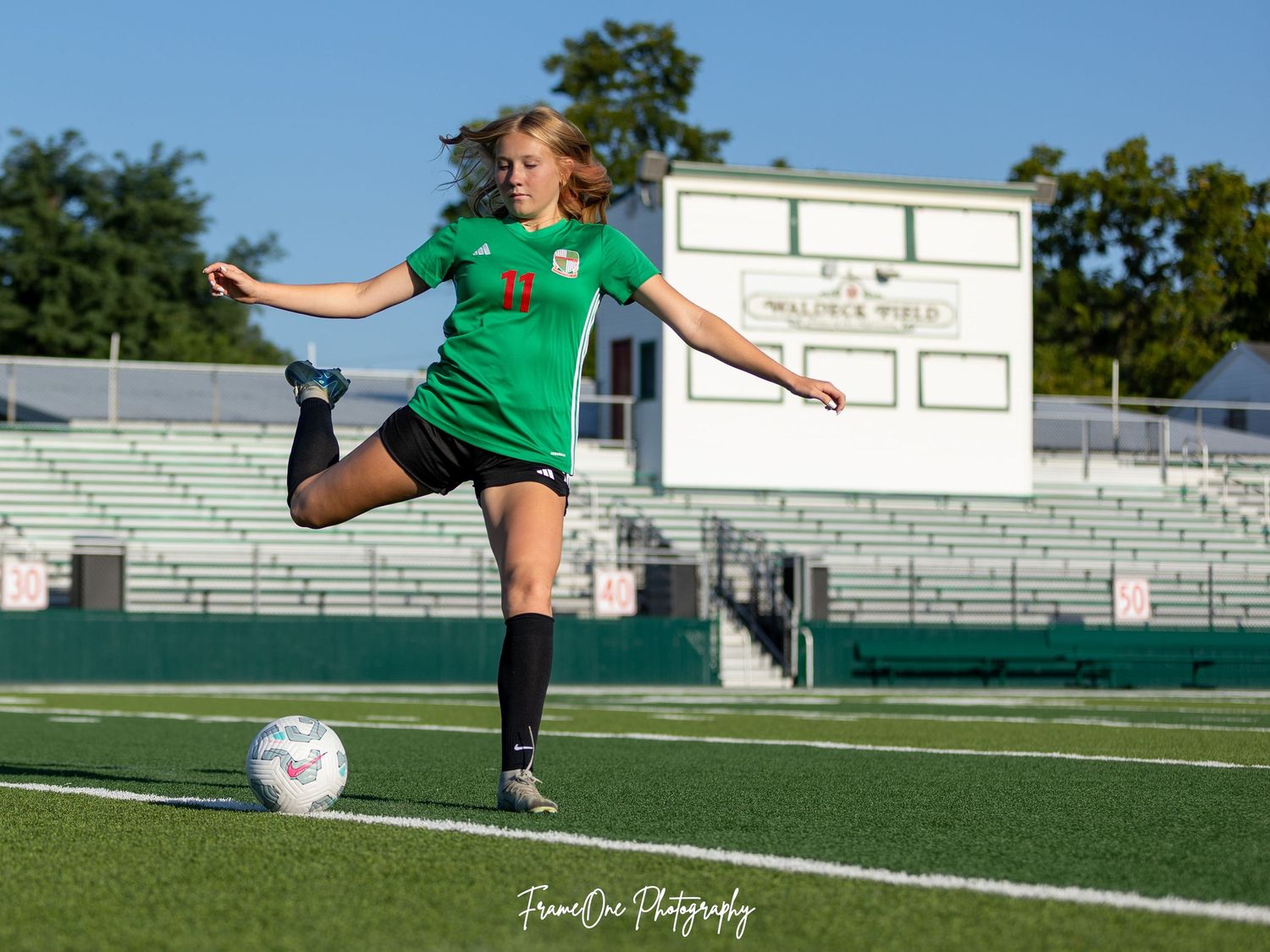 Soccer player in green jersey practices kicking technique on field with stadium bleachers in background.