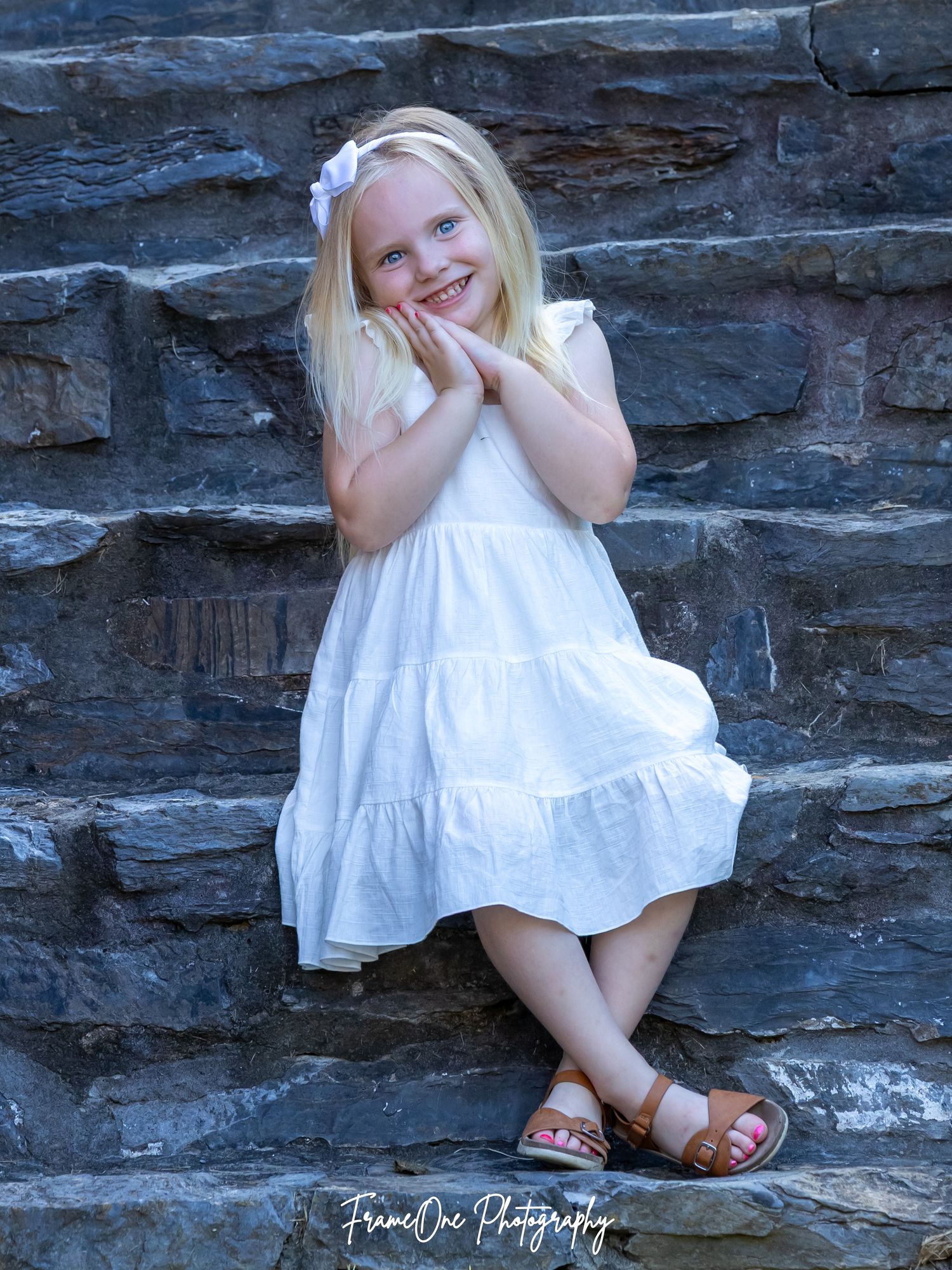 A child in a white summer dress poses playfully against a stone wall wearing brown sandals.
