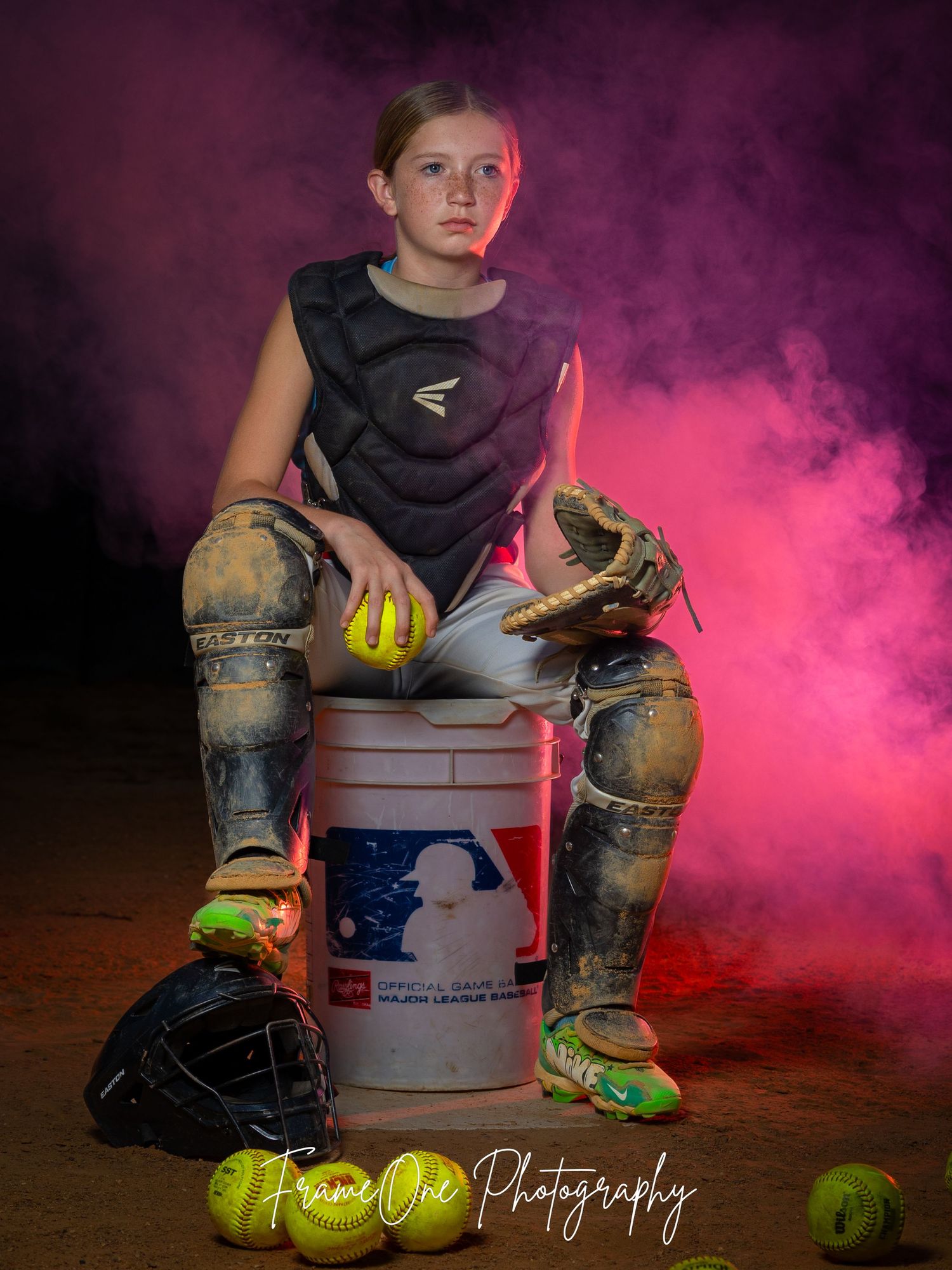 Dramatic portrait of a softball player sitting on MLB bucket surrounded by purple smoke and equipment.