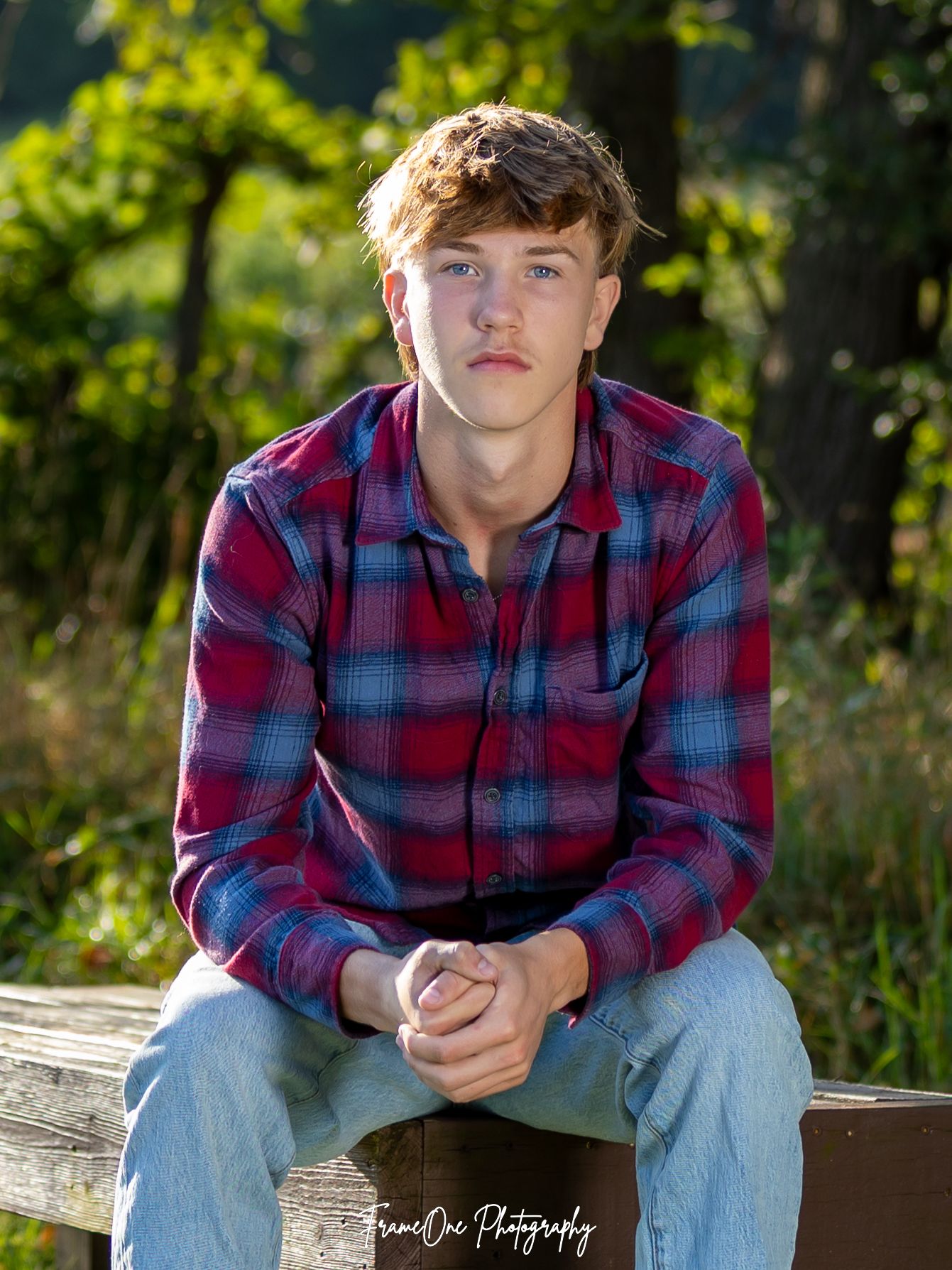Person in a red and blue plaid shirt and jeans sits casually outdoors in a natural setting with green foliage.