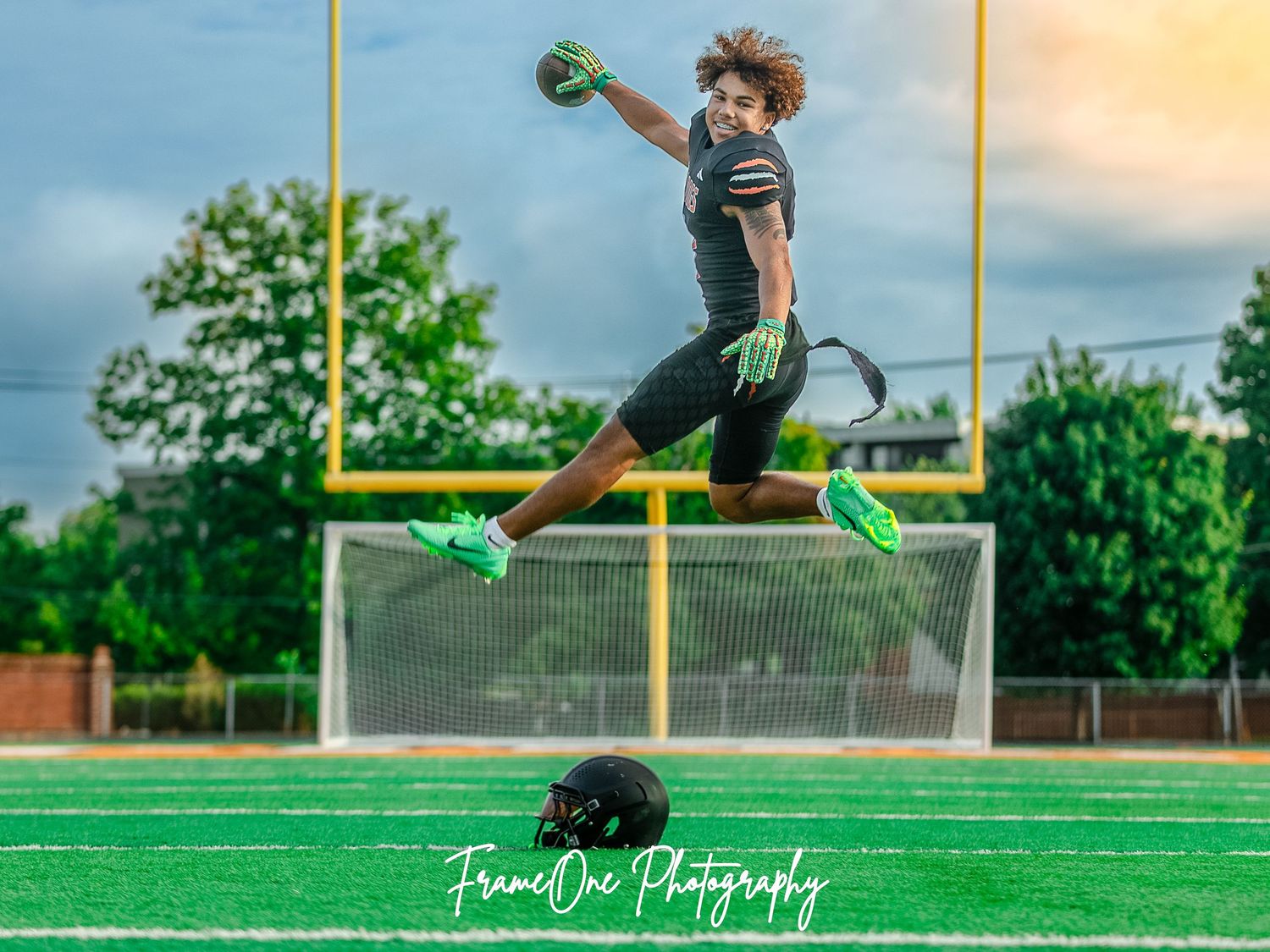 A football player high-steps into the air while jumping towards goalposts on a green field at sunset.