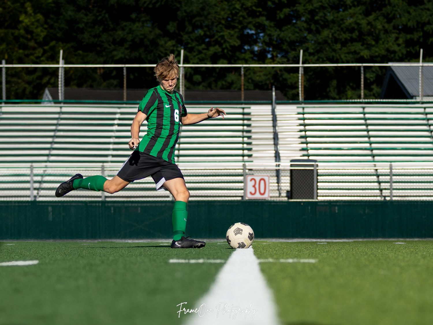 Player in green uniform prepares to kick soccer ball on field with bleachers in background.