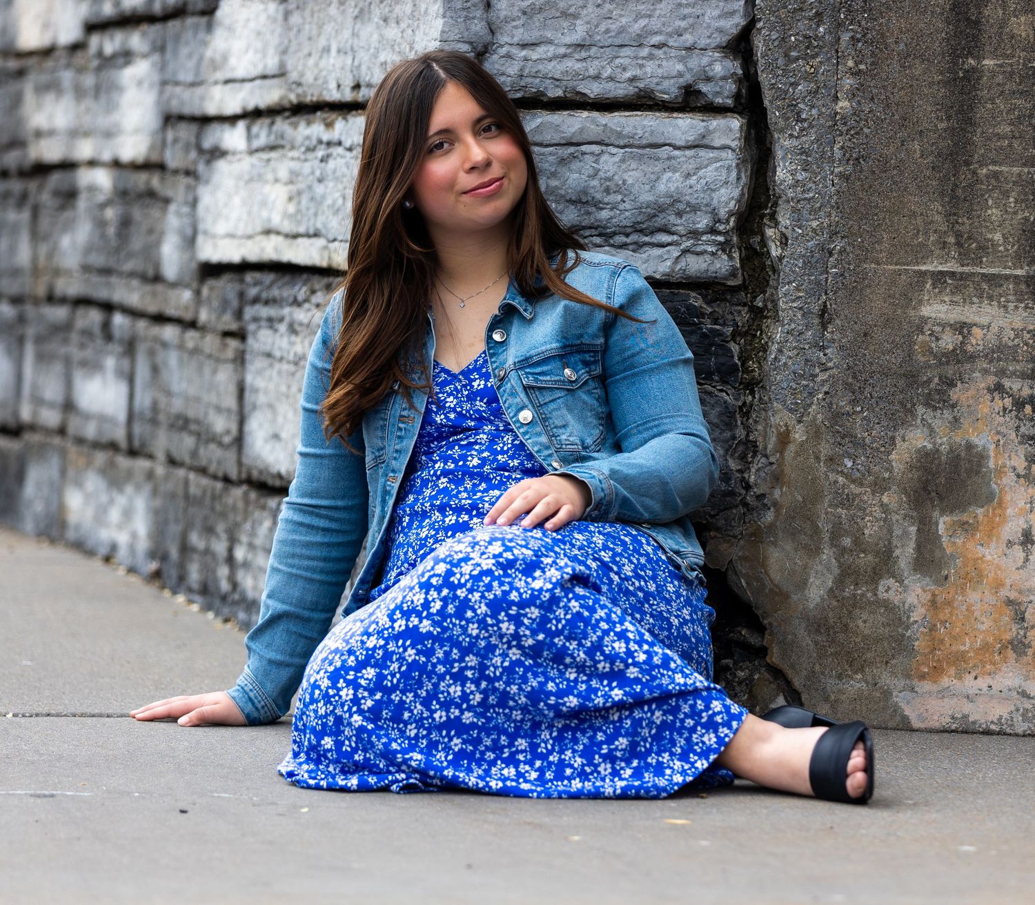 A person in a blue floral dress and denim jacket sits against a stone wall on a city sidewalk.