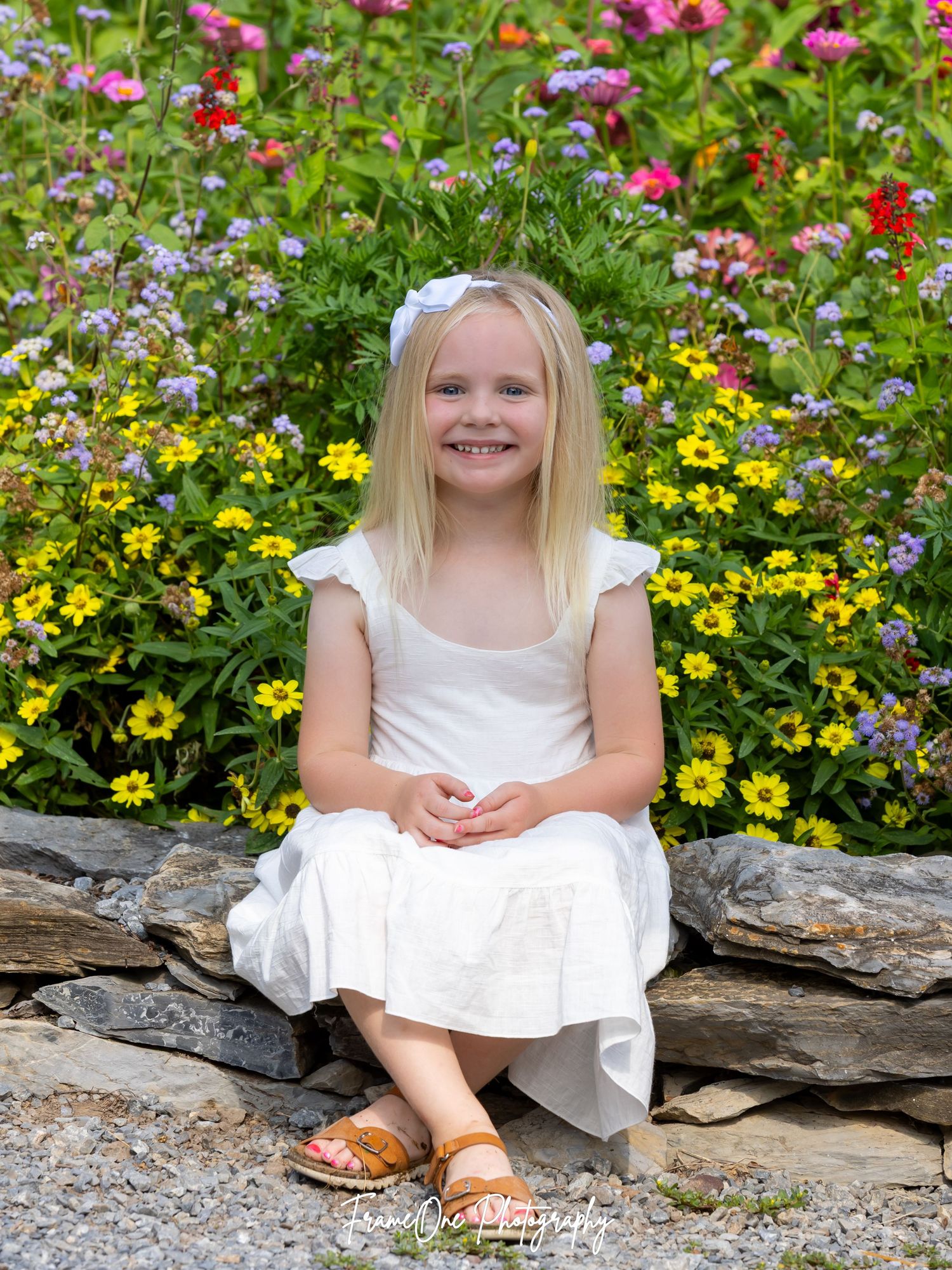 A young person in white dress sits on stone wall surrounded by colorful summer garden flowers.