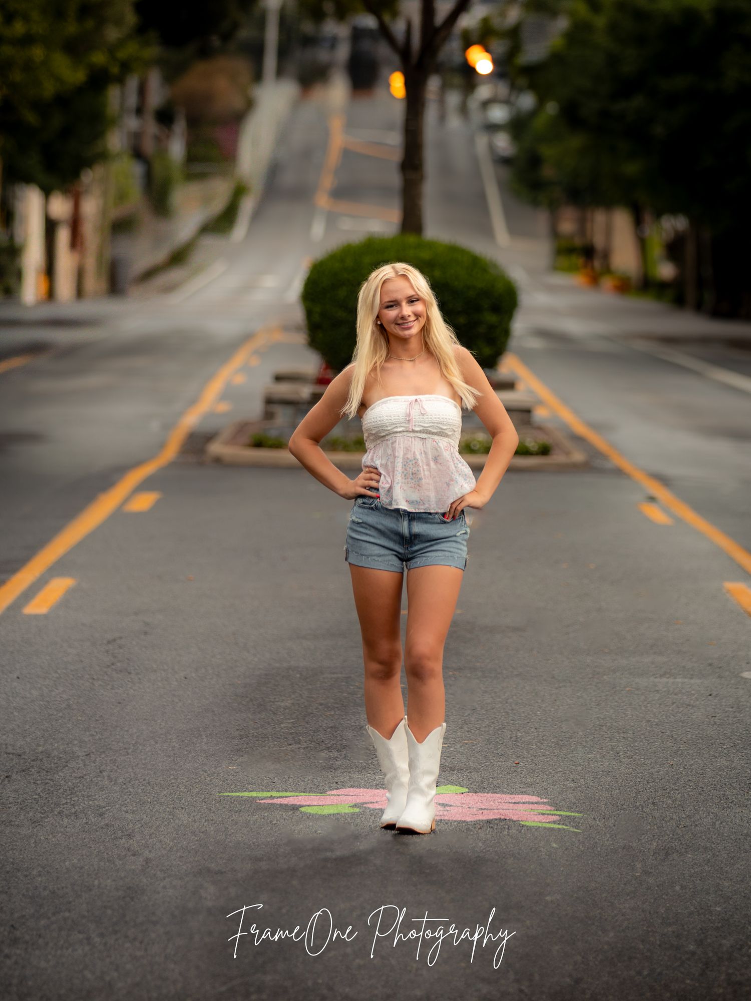 White cowboy boots and denim shorts stand out in a portrait taken on a sloping residential street at dusk.
