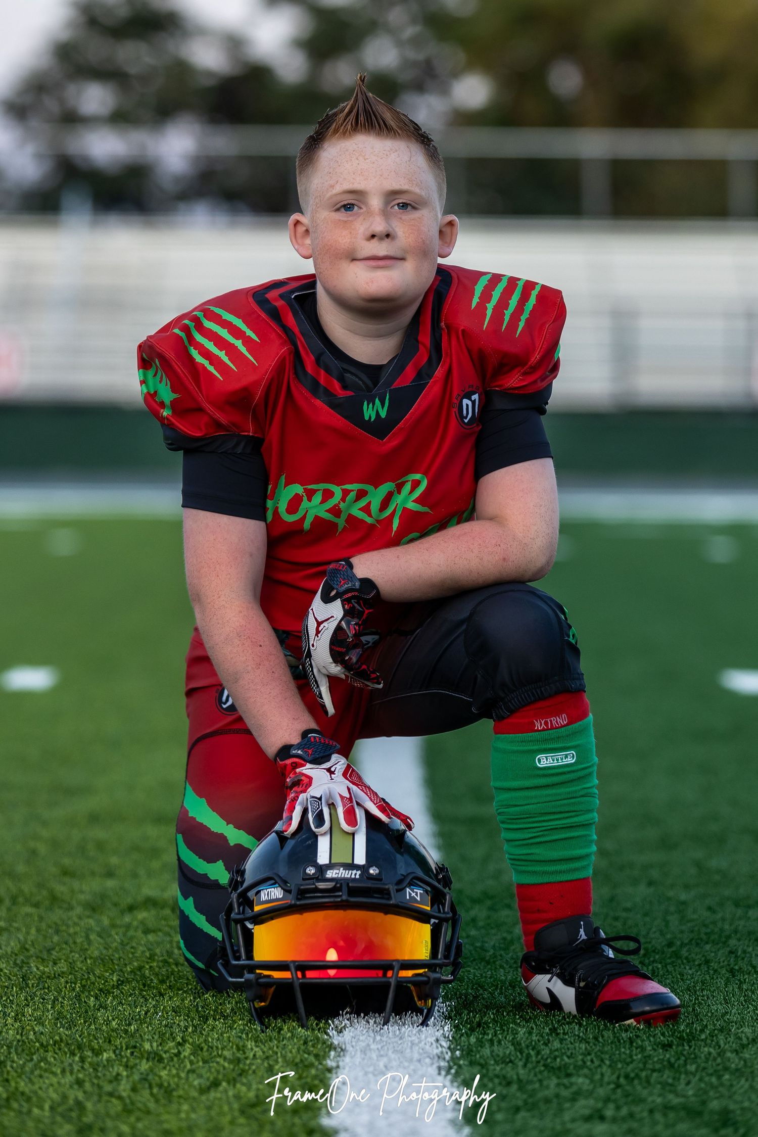 Youth football player sitting on field with helmet, wearing red jersey and black and green uniform in athletic pose.