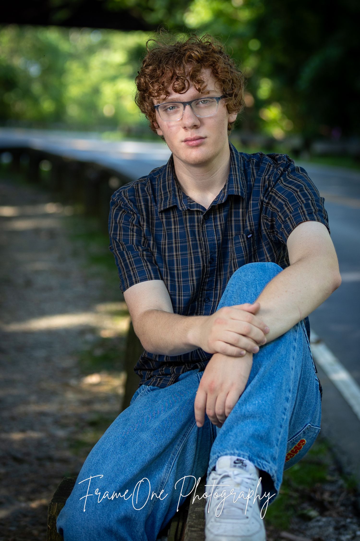 Portrait photo taken outdoors with blurred natural background showing someone in a plaid shirt and jeans sitting on steps.